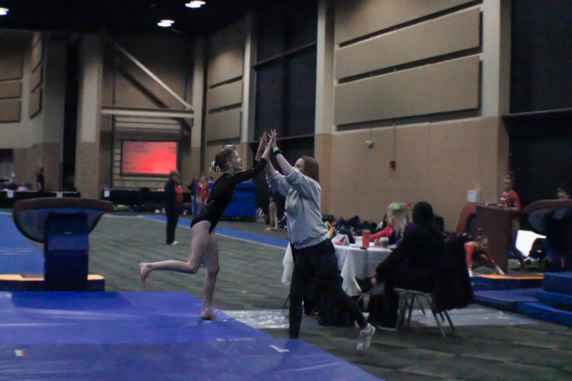 Gymnast leaping for a high-five in an indoor gym, with spectators and blue mats nearby.