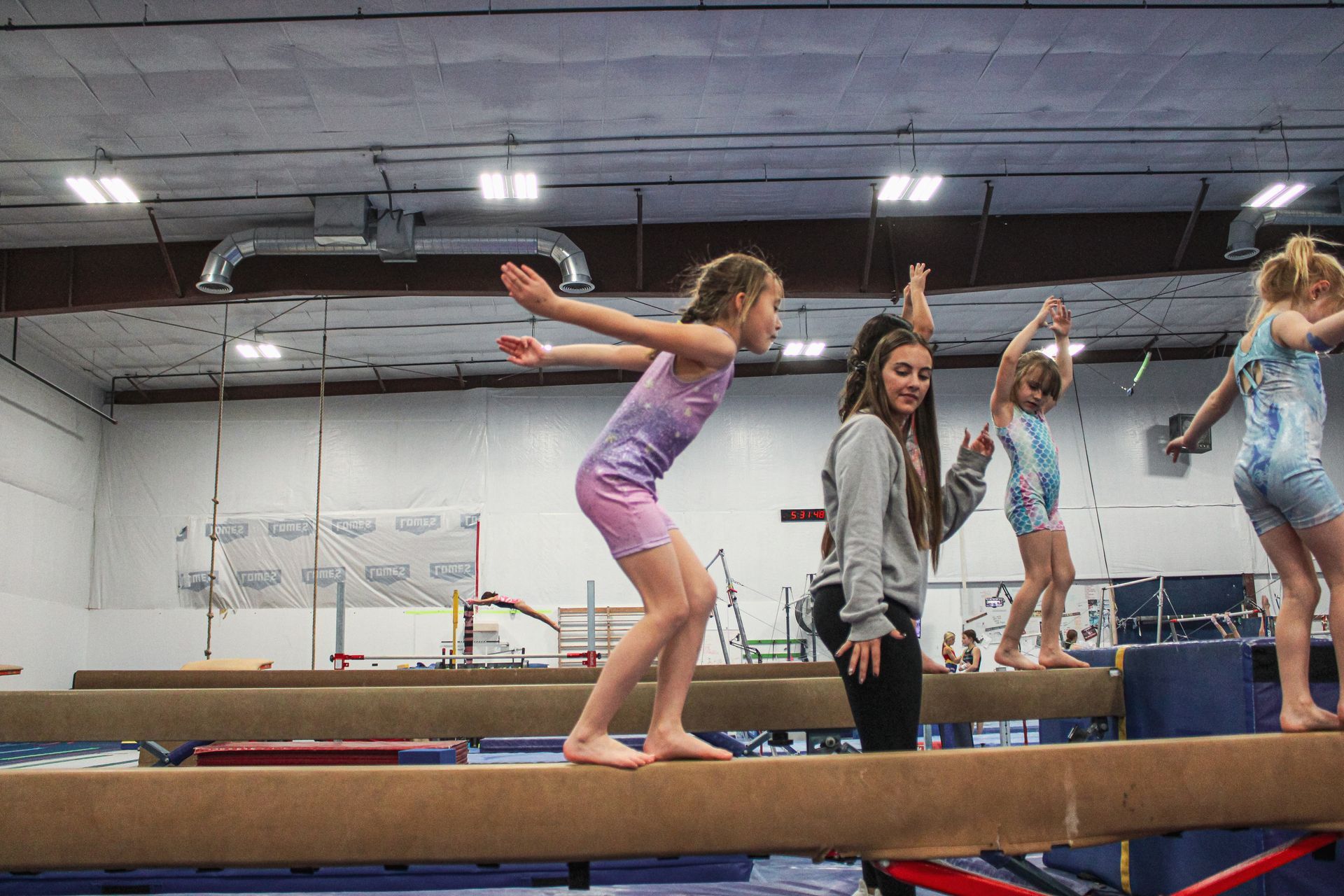 Gymnasts balancing on a beam in a training gym, with a coach standing nearby.