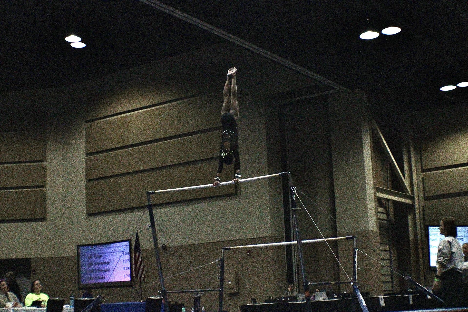 Gymnast balancing upside down on a high bar in a dim indoor arena