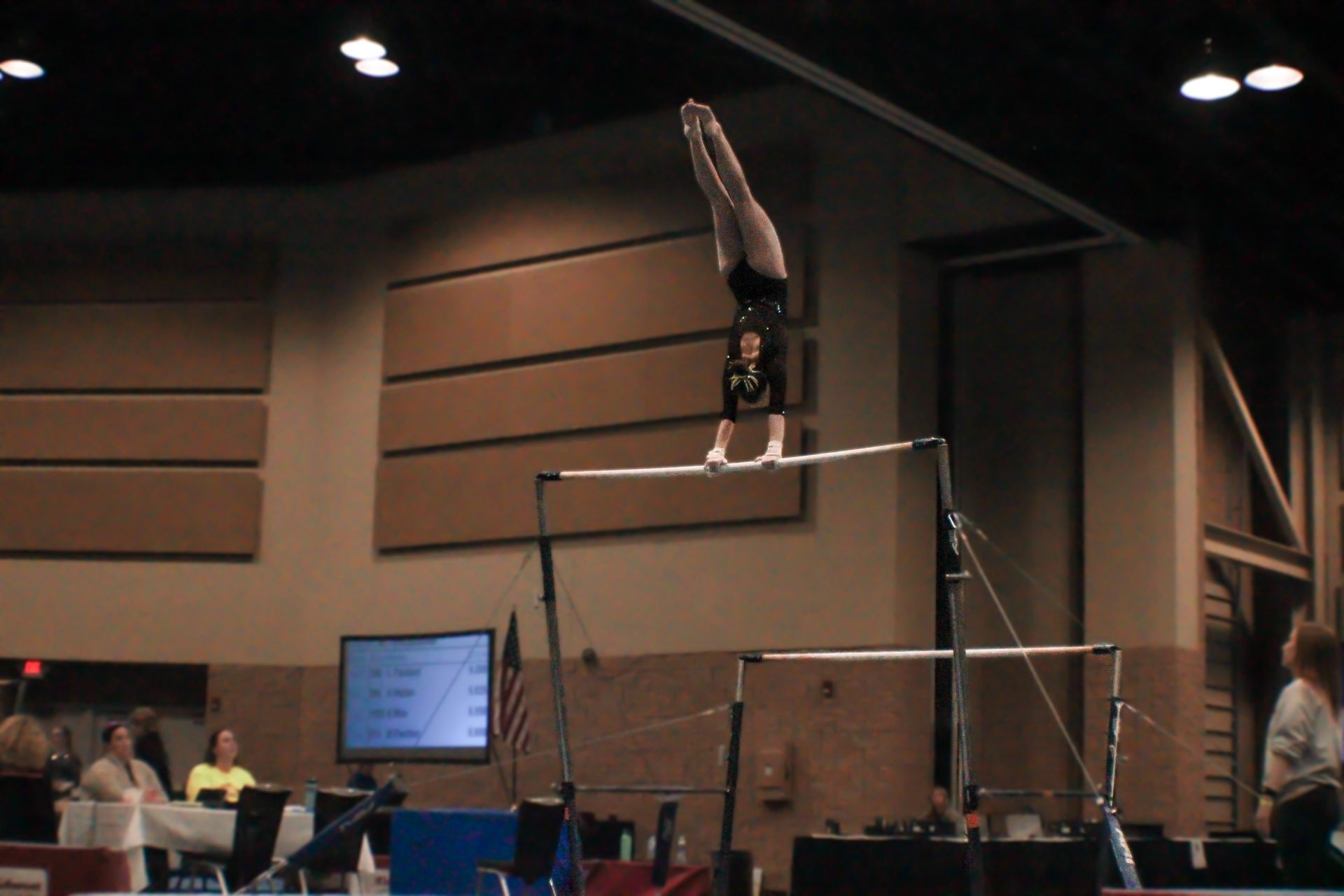 Gymnast upside down on uneven bars in an indoor competition arena, with judges and spectators nearby