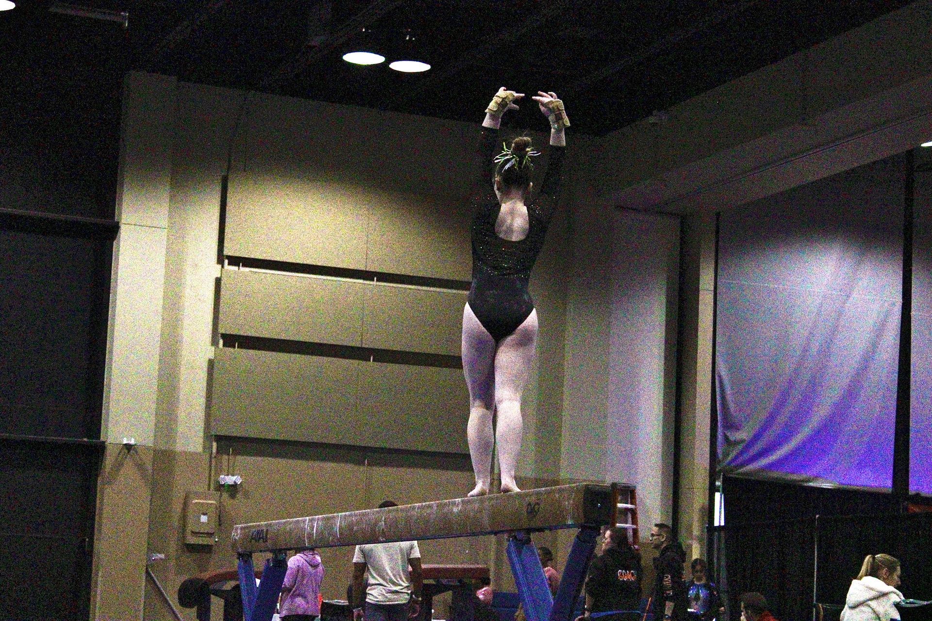 Gymnast balancing on a beam with arms raised in an indoor competition venue