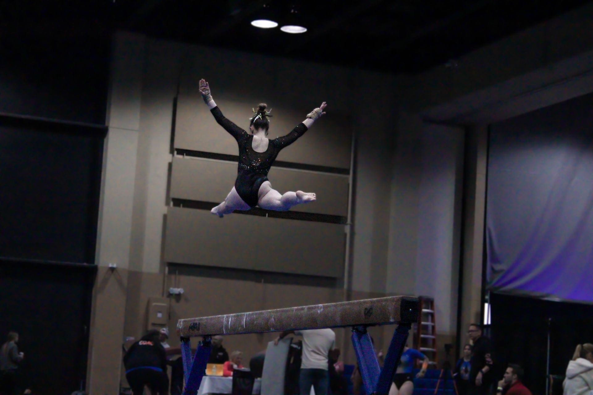 Gymnast midair above a vault table, arms raised in a dark indoor arena.