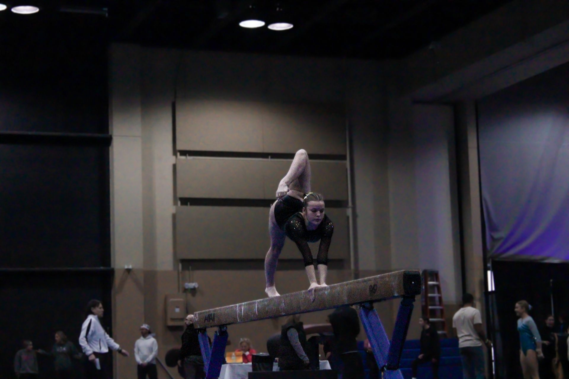 Gymnast balancing on a raised beam in a dim indoor auditorium, with spectators below