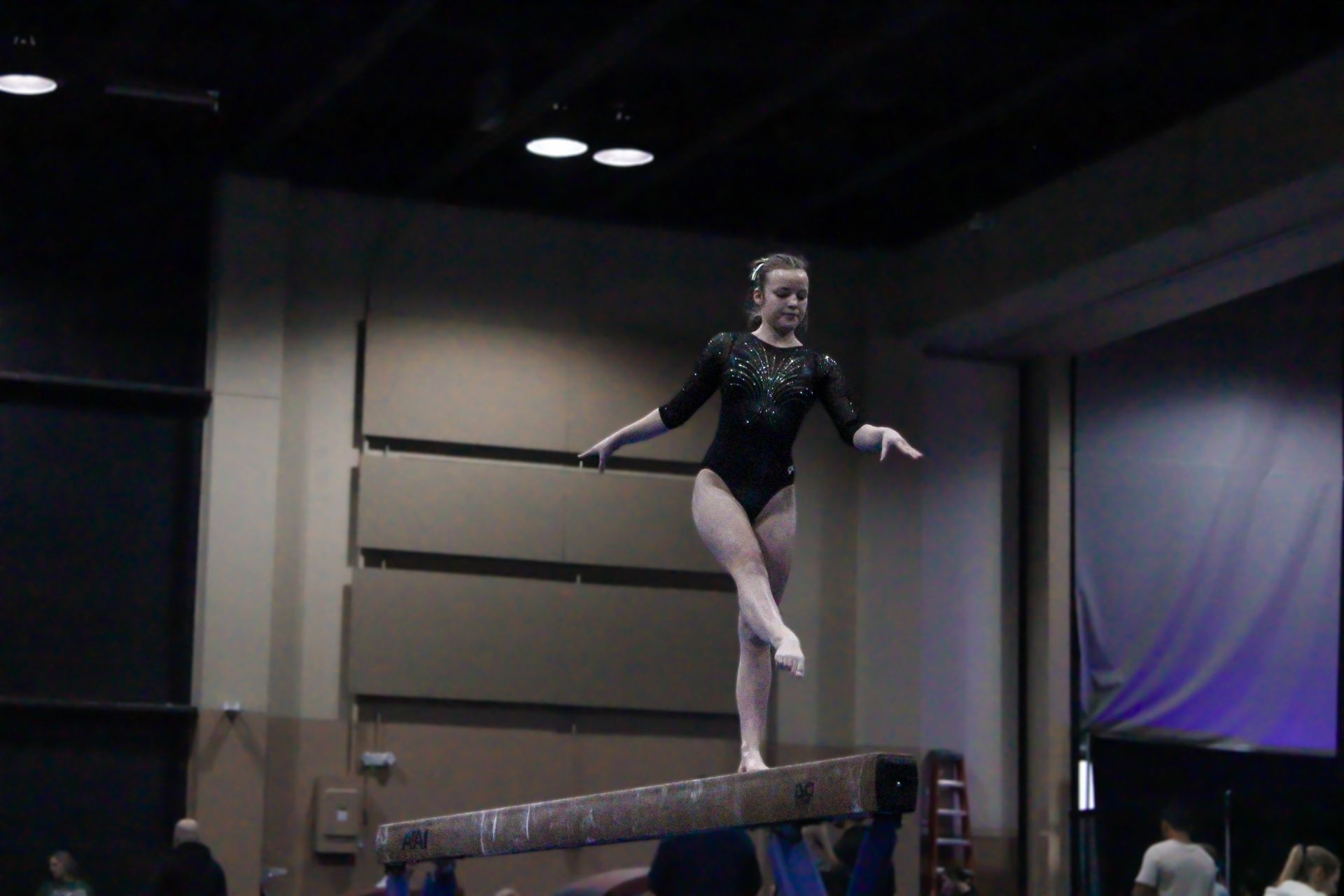 Gymnast balancing on a beam in a dim indoor gym, wearing a black leotard.