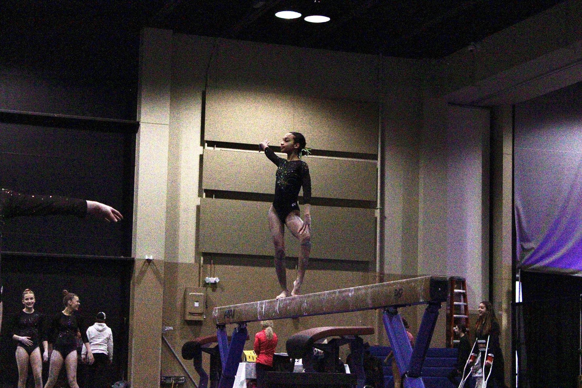 Gymnast balancing on a high beam indoors, with spectators watching below.
