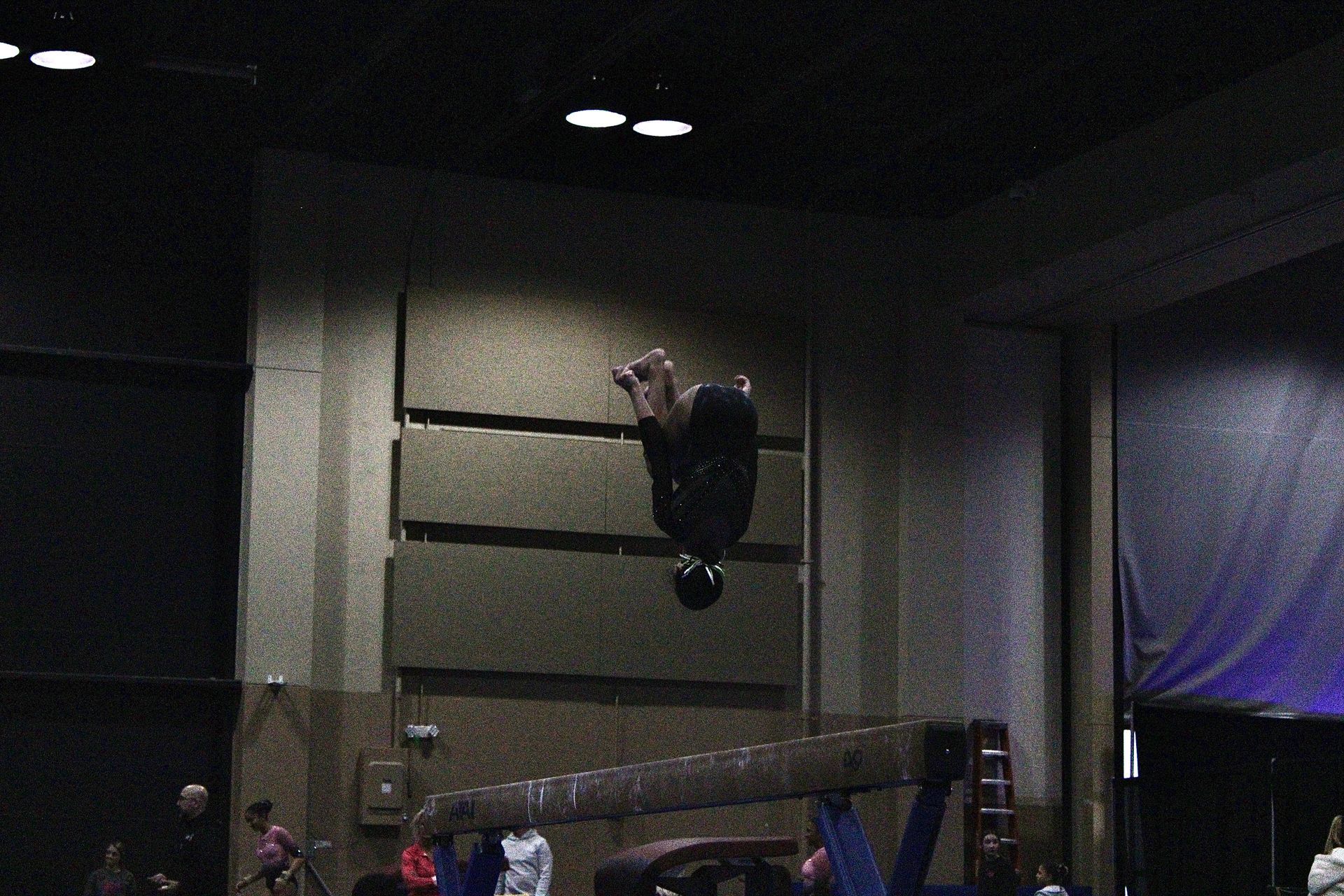 Person hanging upside down from a high wall in a dim indoor gym, with spectators below