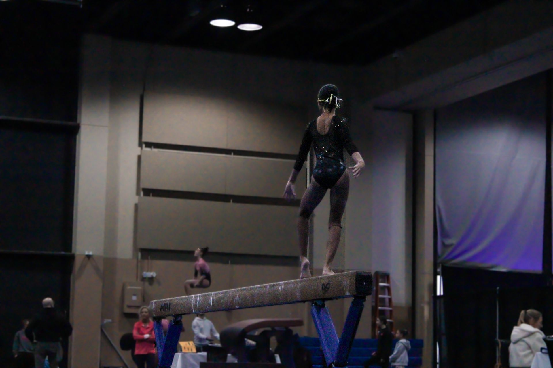 Gymnast balancing on a beam during a competition, with judges and spectators in the background.