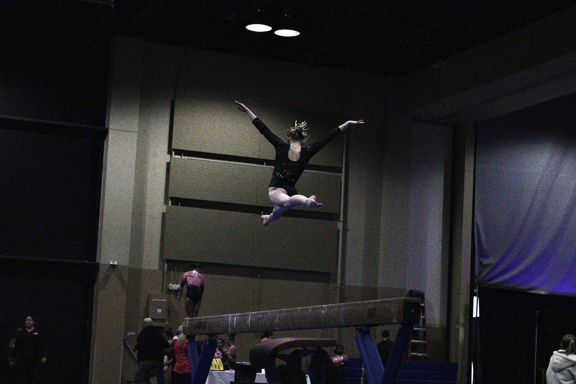 Gymnast midair above a trampoline in a dark indoor training facility, arms and legs spread wide.