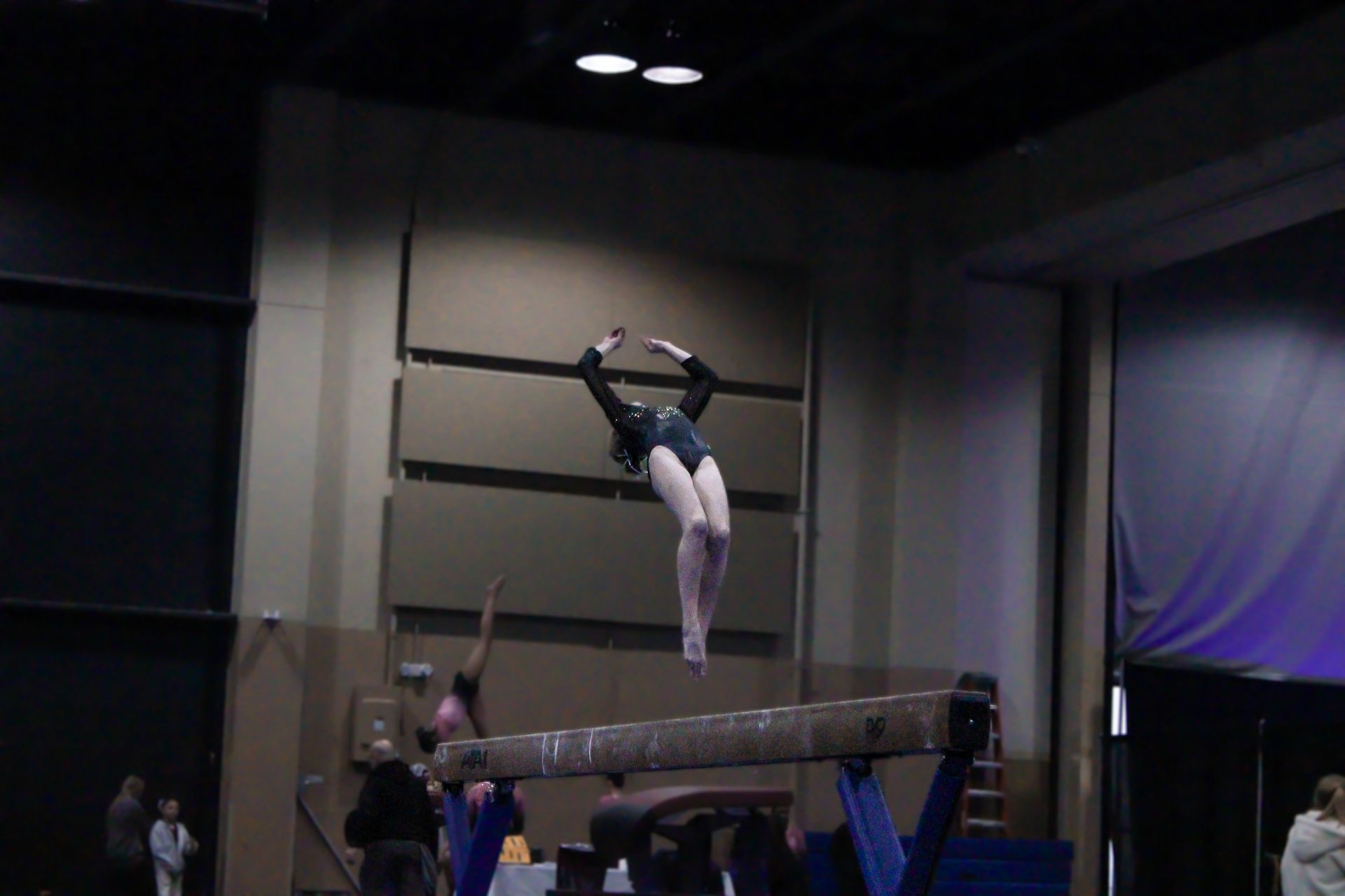 Gymnast performing a leap above the balance beam in an indoor competition arena.
