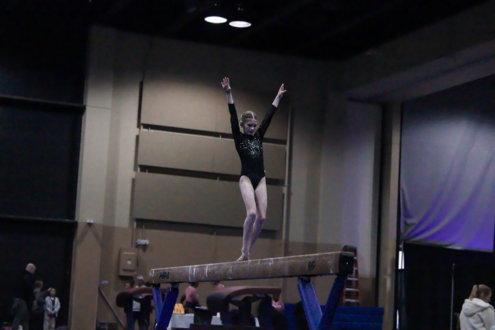 Gymnast standing on a balance beam with arms raised in a dim indoor gymnasium