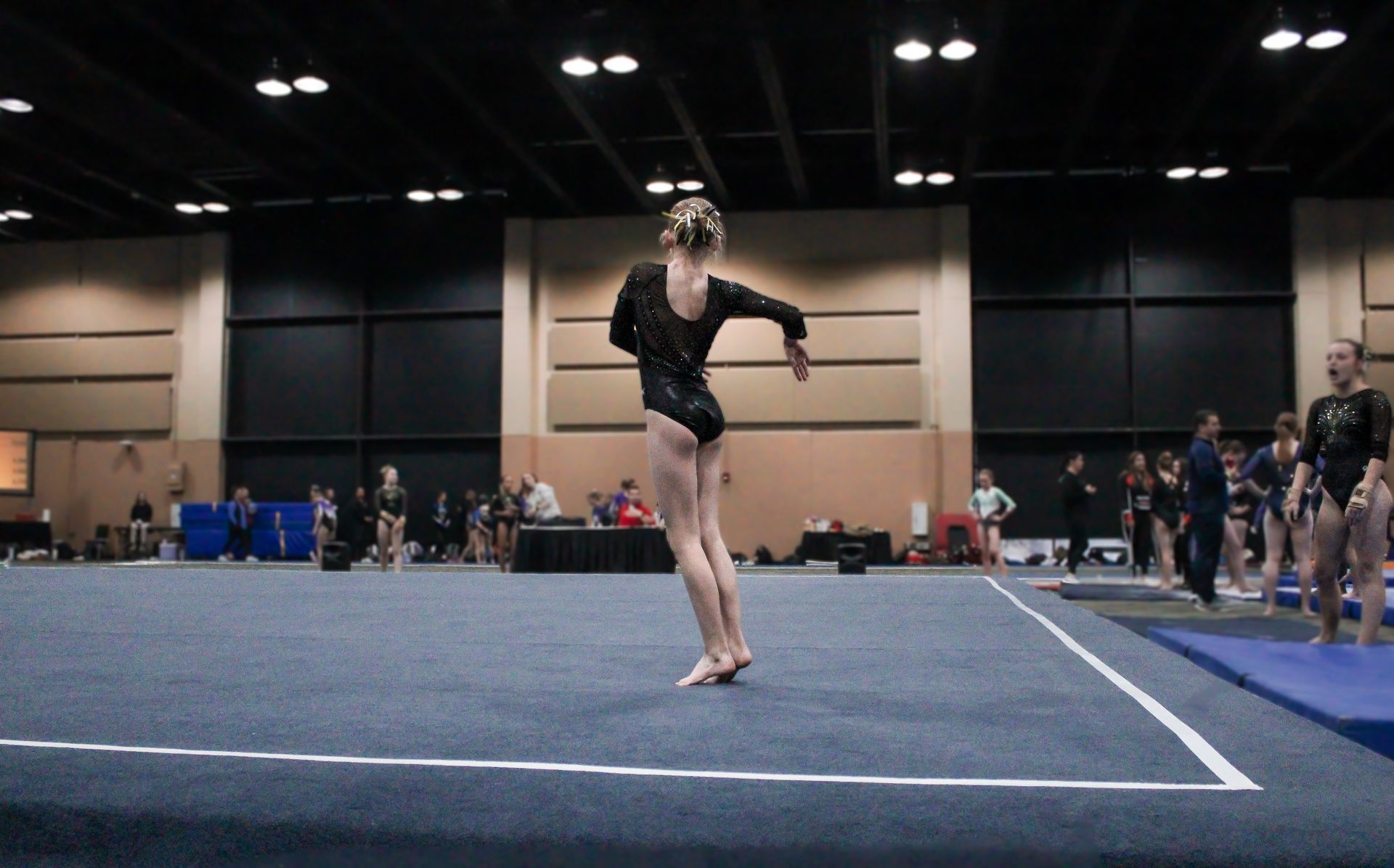 Gymnast balancing on one foot on a blue mat in an indoor competition arena.