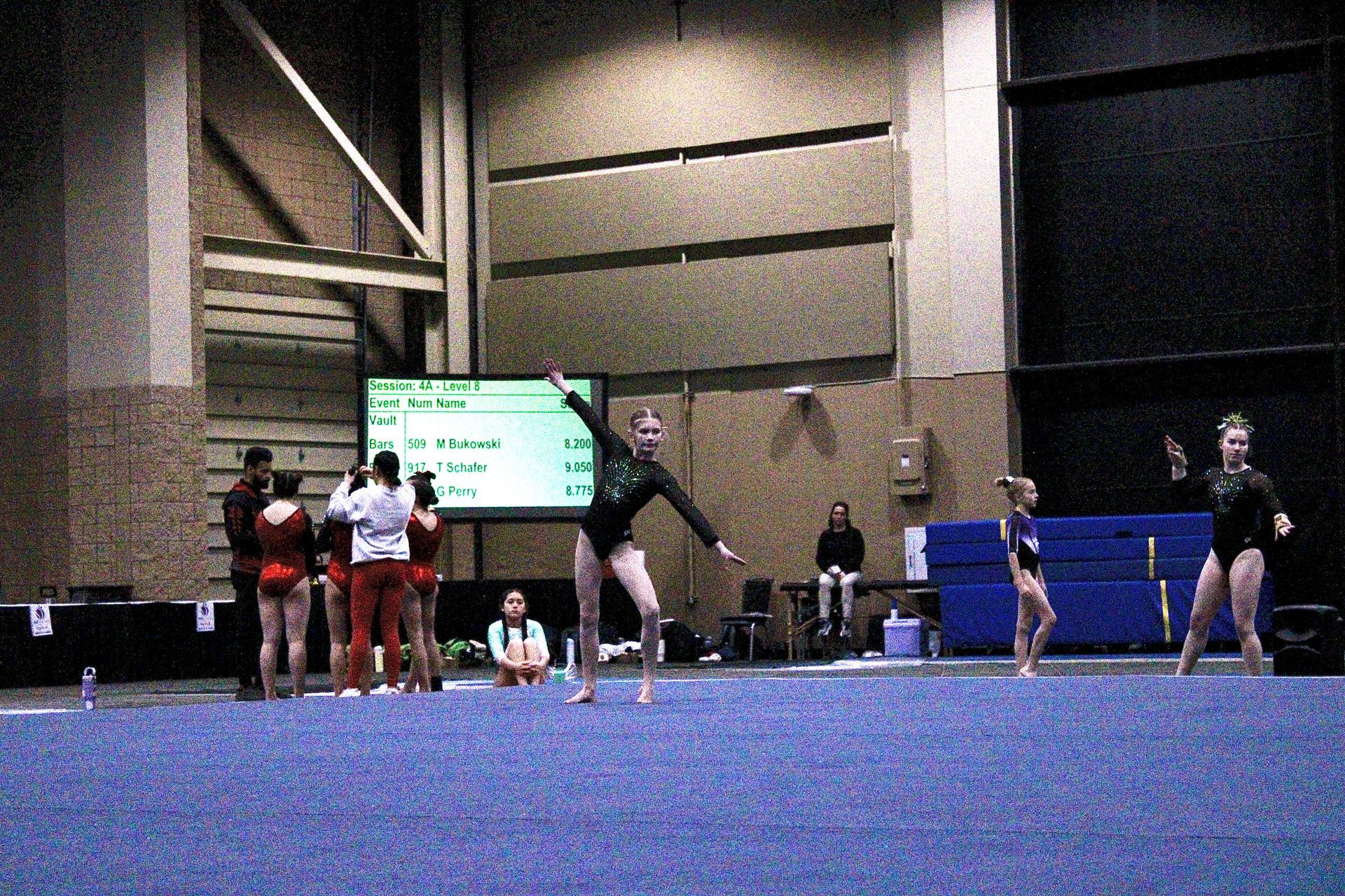 Gymnast performing a leap on a blue floor mat with teammates and judges in the background