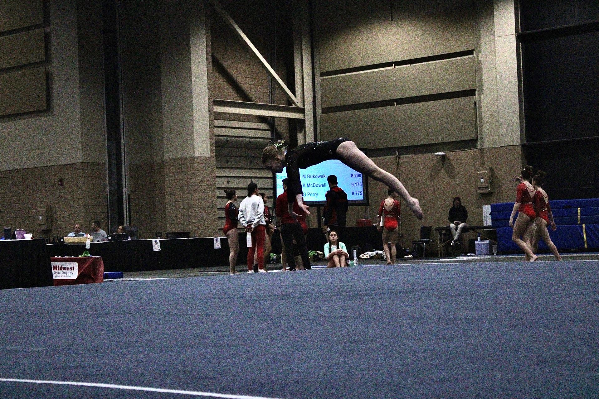Gymnast midair on a blue mat in an indoor arena, with spectators and coaches in the background