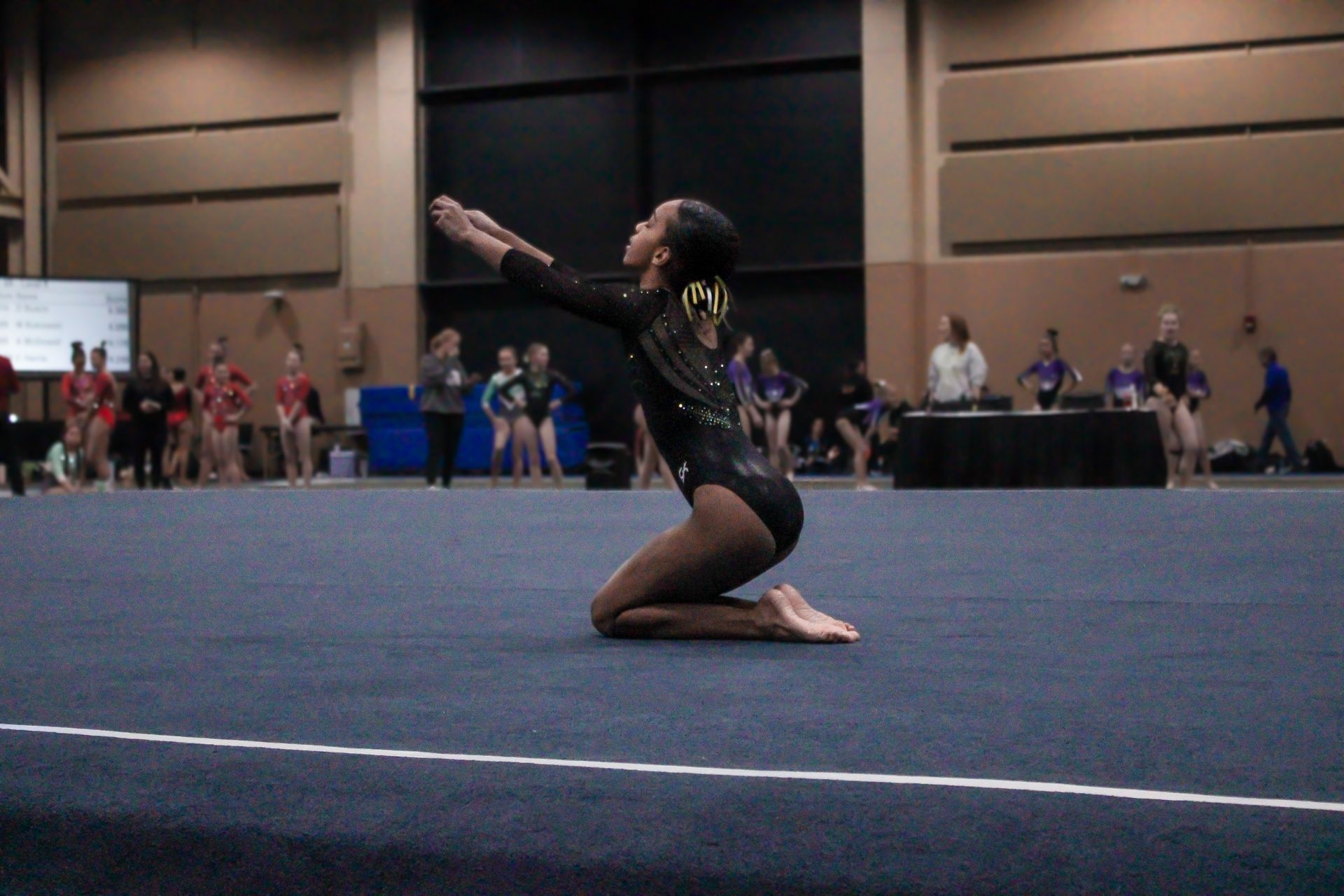 Gymnast kneeling on a blue mat, arms raised during a floor routine in an indoor arena