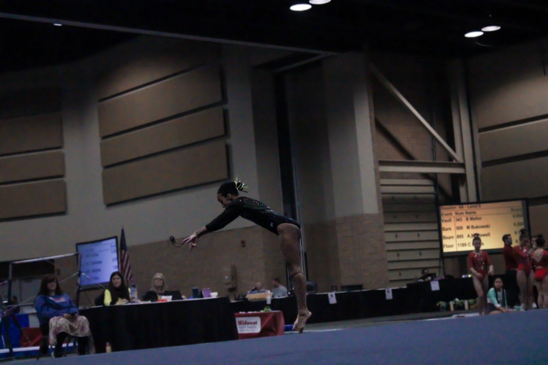 Gymnast balancing on a beam in a large indoor competition hall, arms extended for balance.