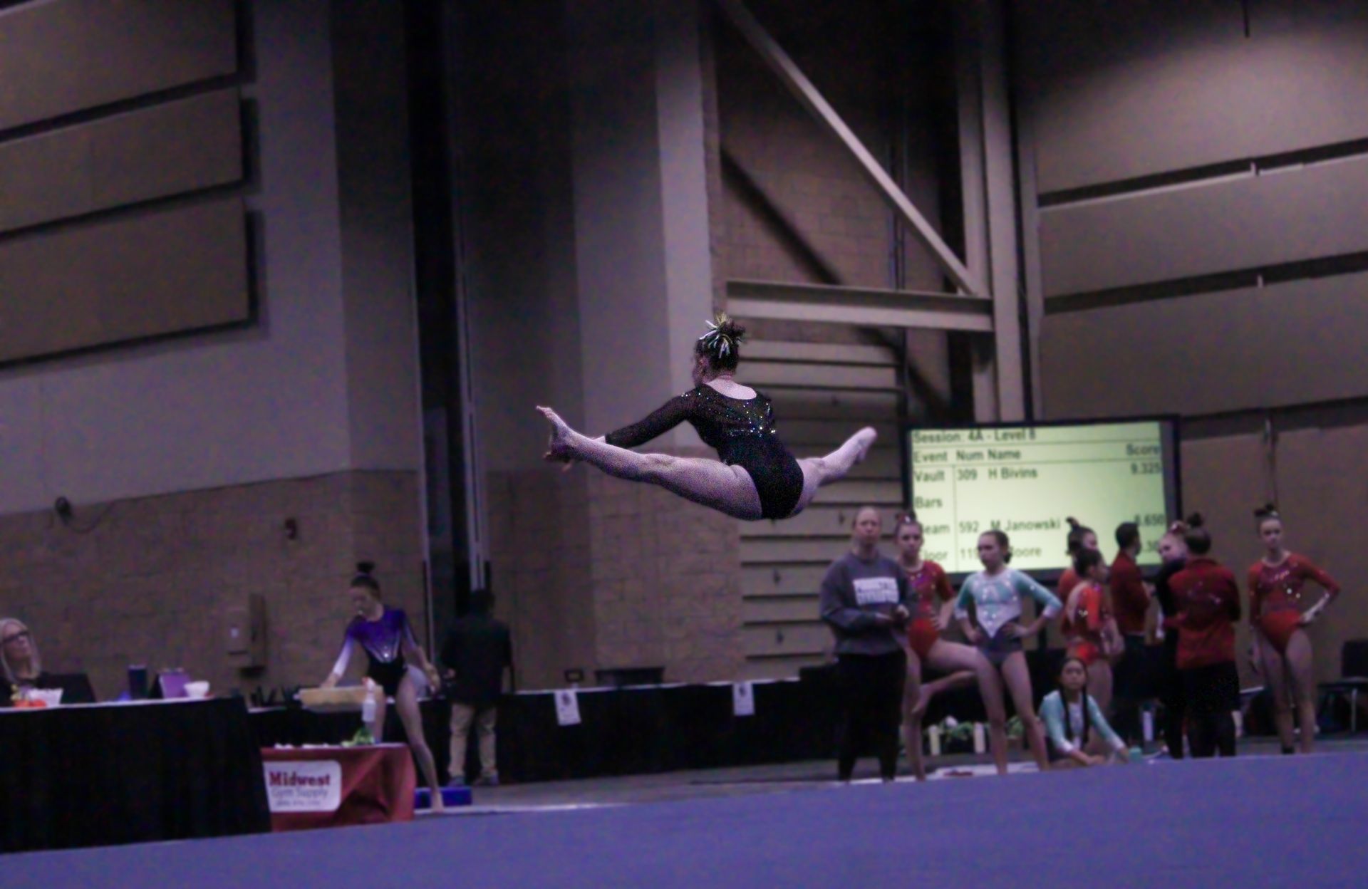 Gymnast midair in a split leap at an indoor competition, with judges and spectators in the background.