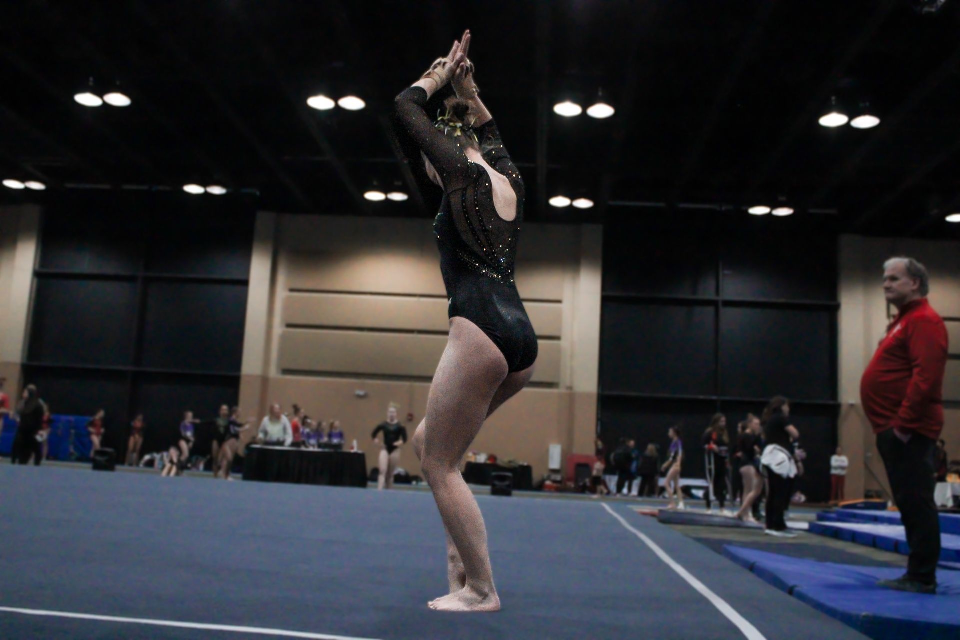 Gymnast performing a floor routine in a gymnasium, arms raised with judges and spectators in the background