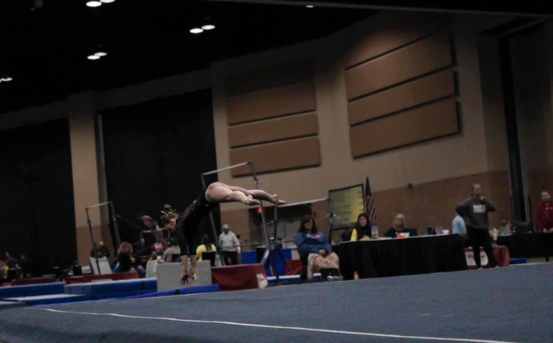 Gymnast mid-air during a vault routine in an indoor competition gym, with judges and spectators in the background.