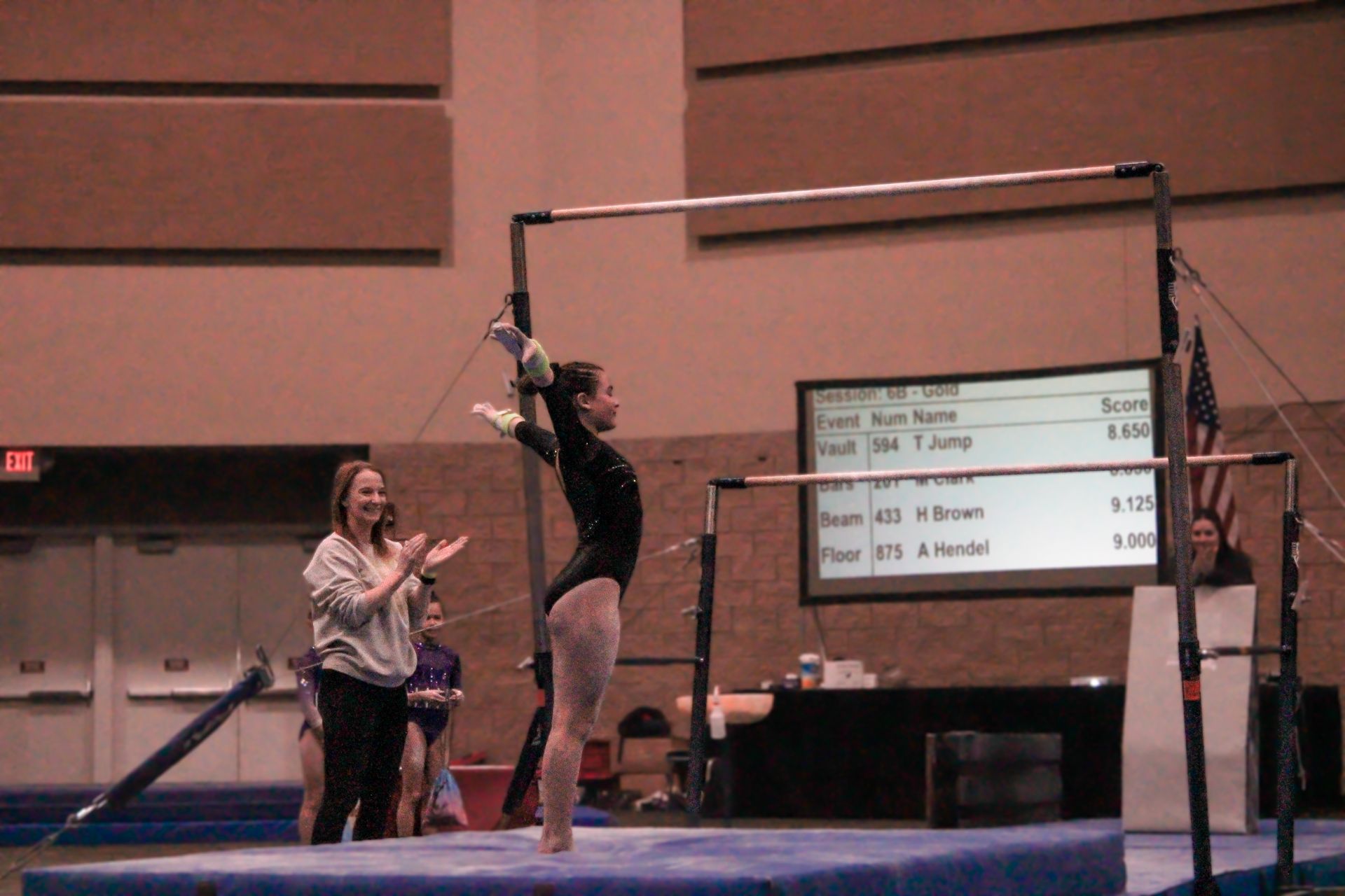 Athlete swings on uneven bars during a gymnastics meet while a coach stands nearby.