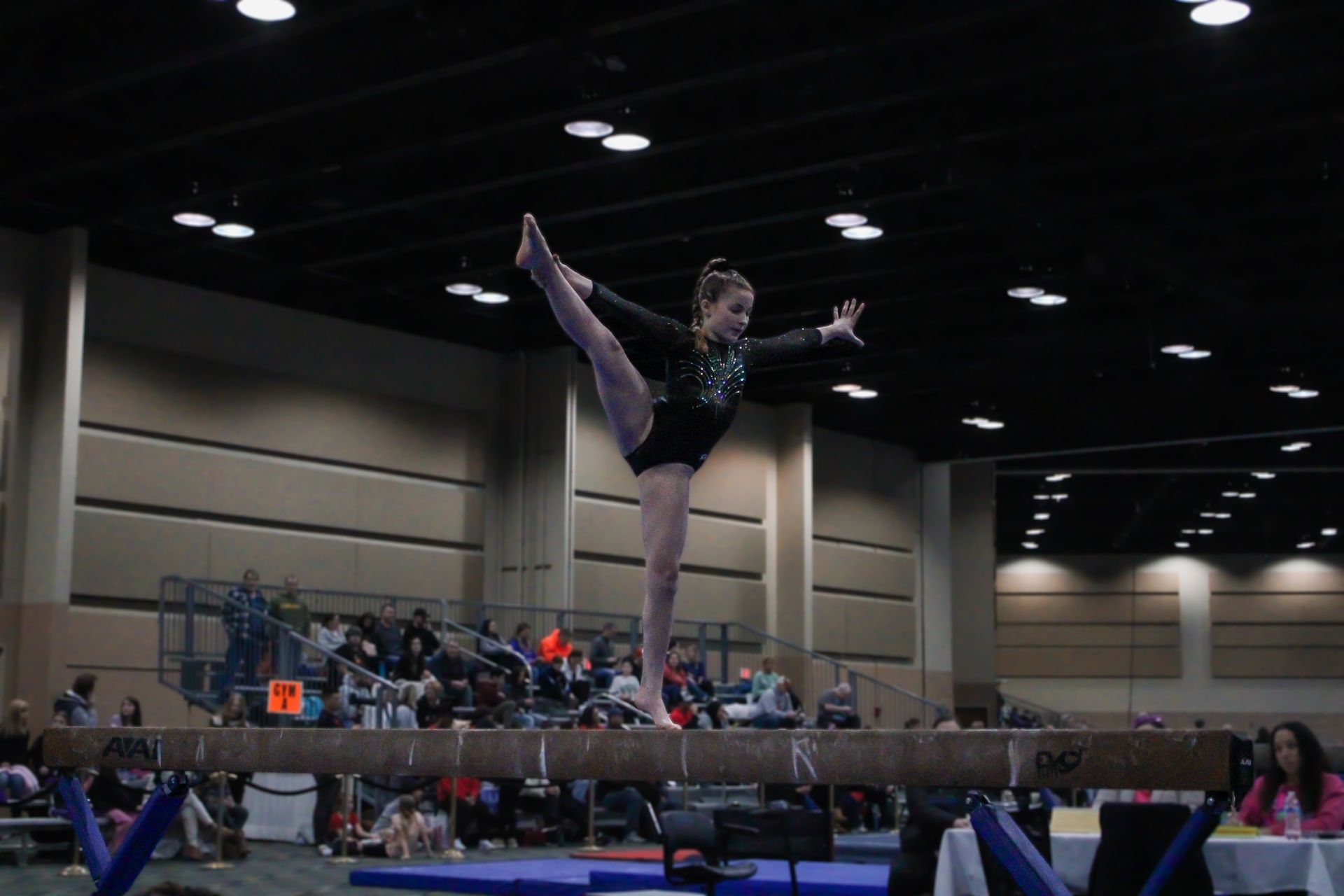Gymnast balancing on one leg on a beam in a large indoor competition arena