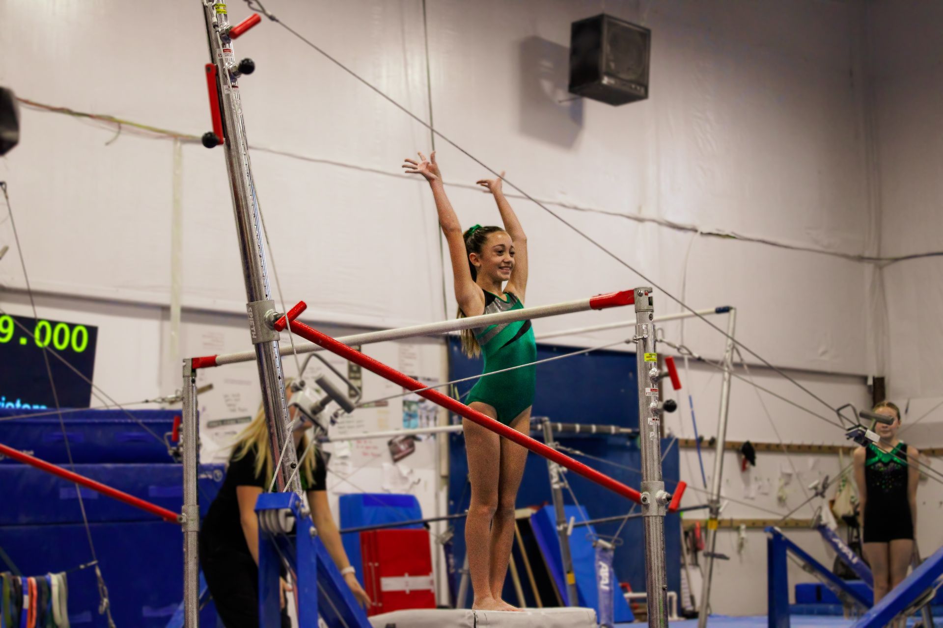 Gymnast in green leotard on uneven bars in an indoor gym, reaching high during a routine