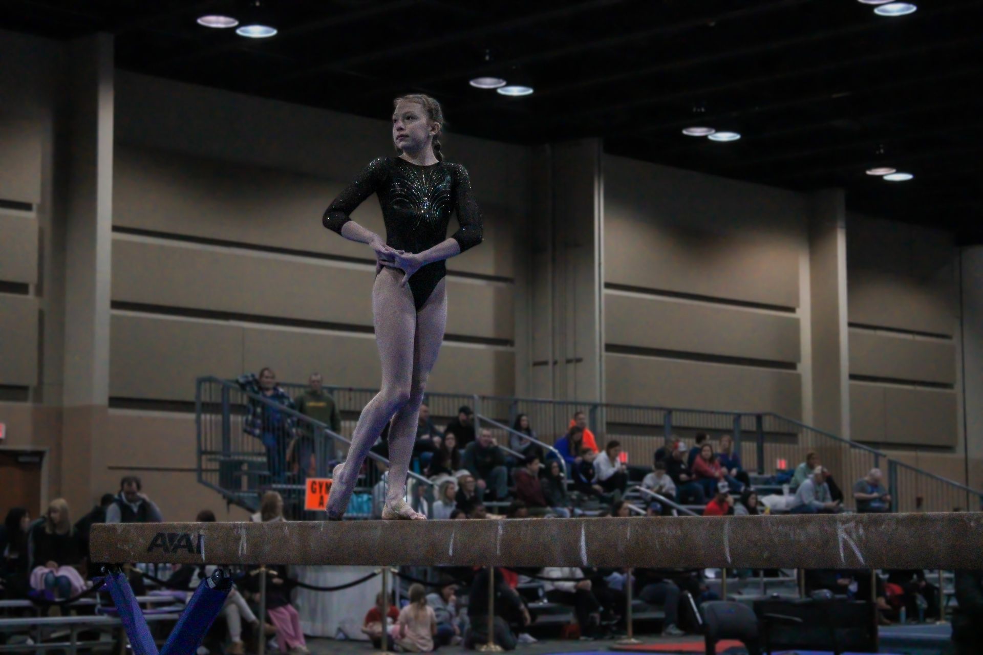 Gymnast in a black leotard balancing on a beam at an indoor competition, with spectators in the background