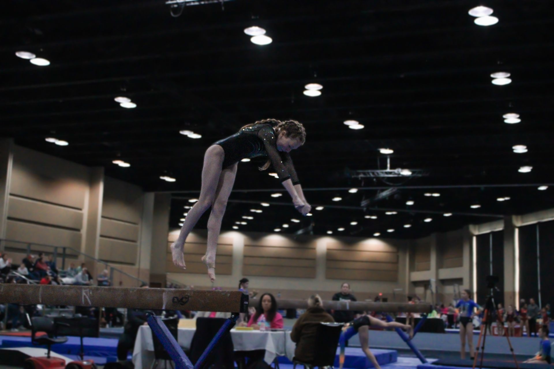 Gymnast mid-flip on a trampoline in an indoor gymnasium, with spectators in the background