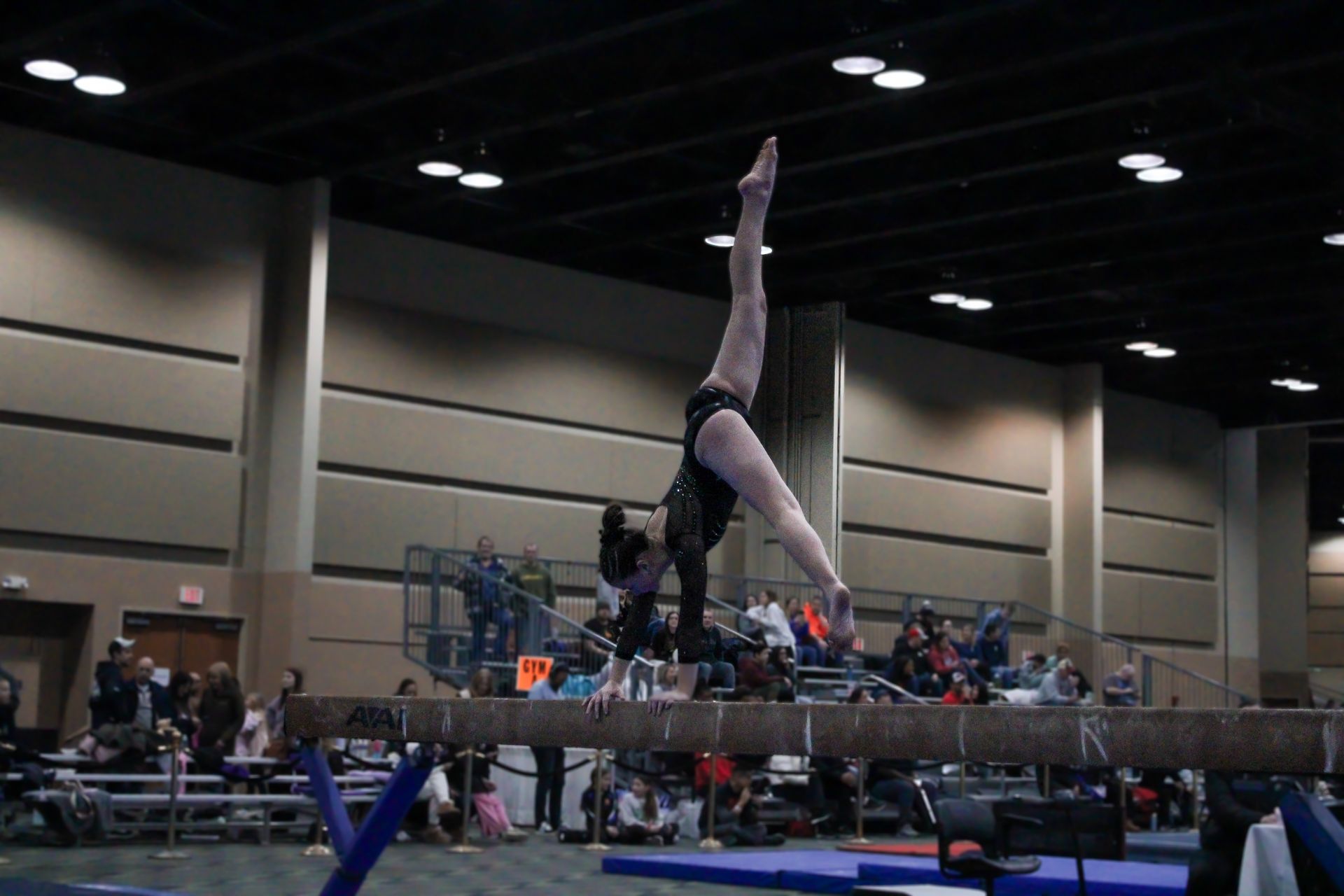 Gymnast balanced upside down on a beam at an indoor competition, with spectators in the background