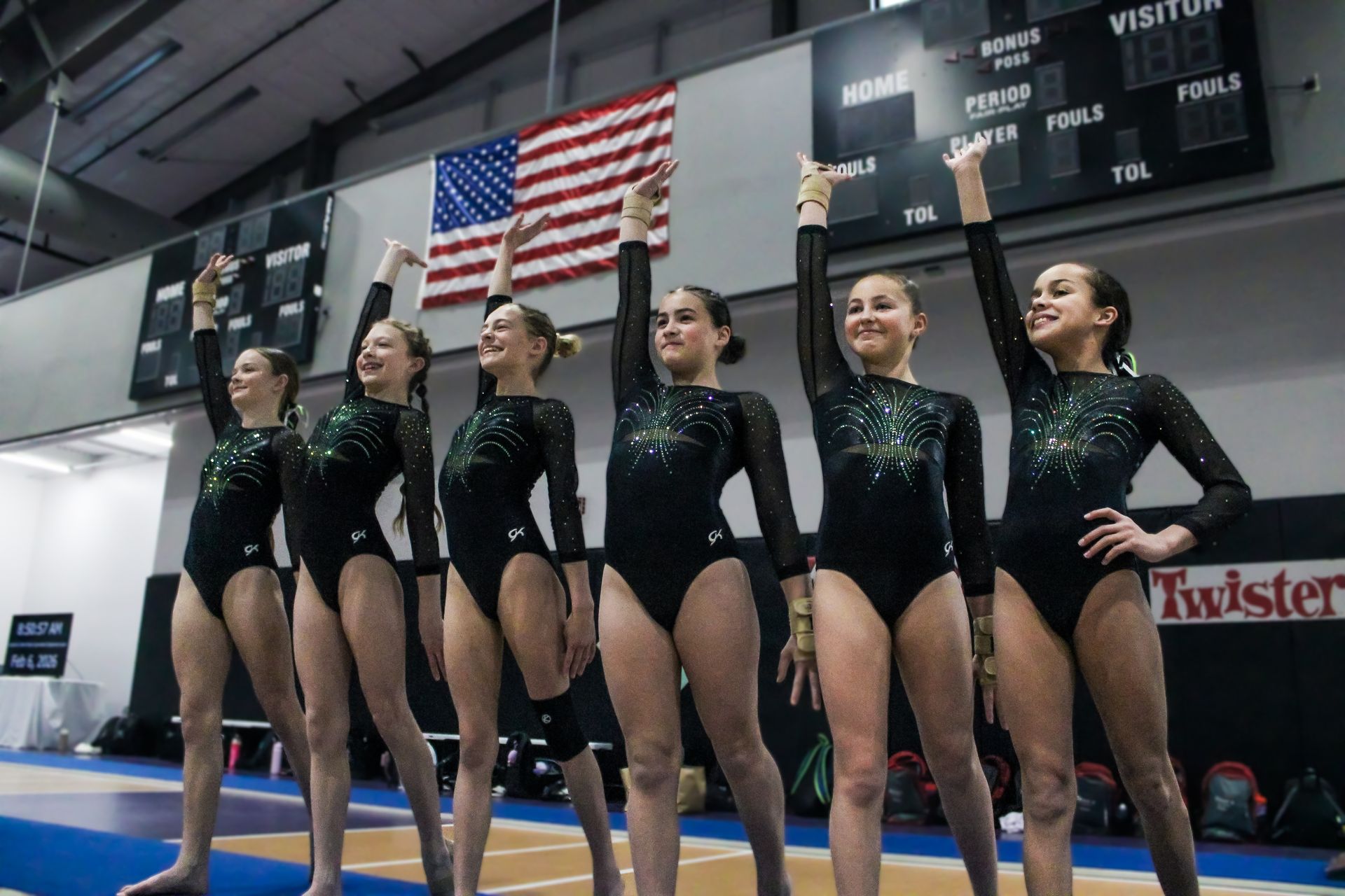 Six gymnasts in black leotards stand lined up on a gymnastics floor beneath an American flag.
