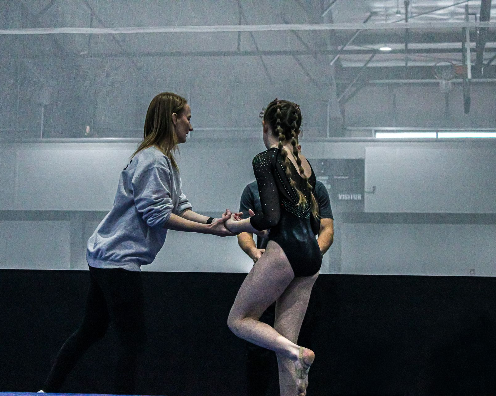 Two women holding hands by an indoor gymnastics mat, one in a black leotard standing on tiptoe.
