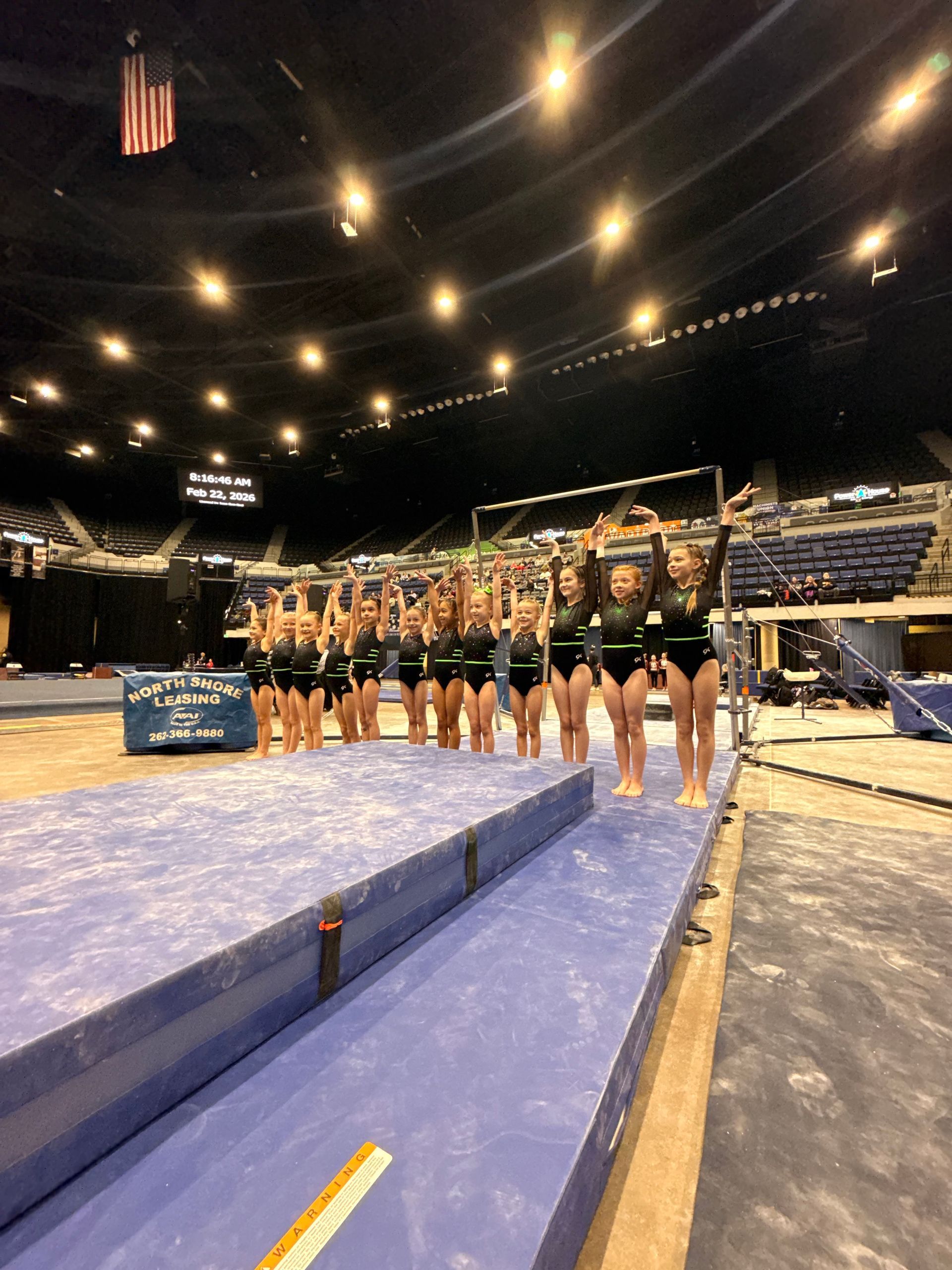 Gymnasts lined up on a blue vault runway inside a dimly lit indoor arena.