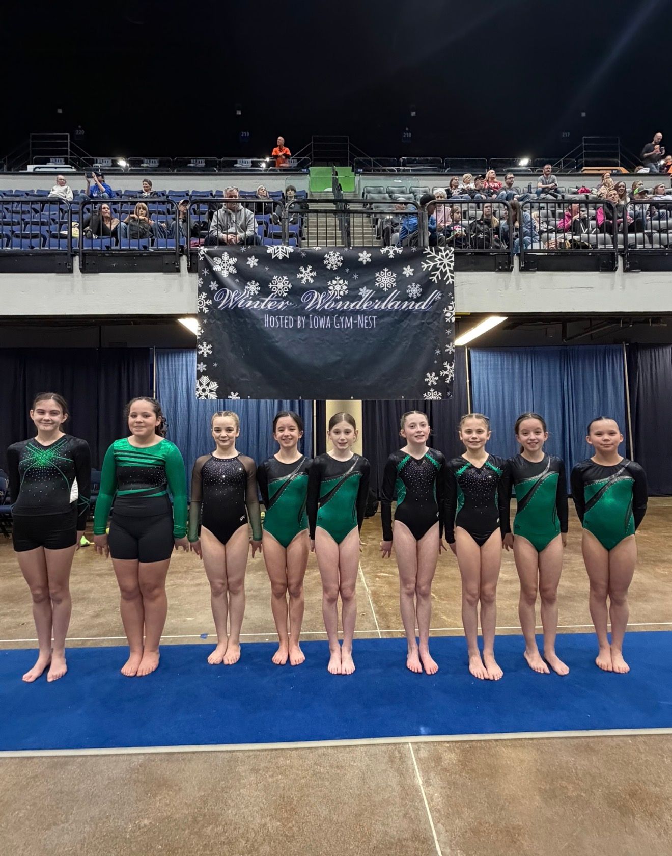Gymnastics team in green leotards standing on a blue mat at an indoor arena awards ceremony