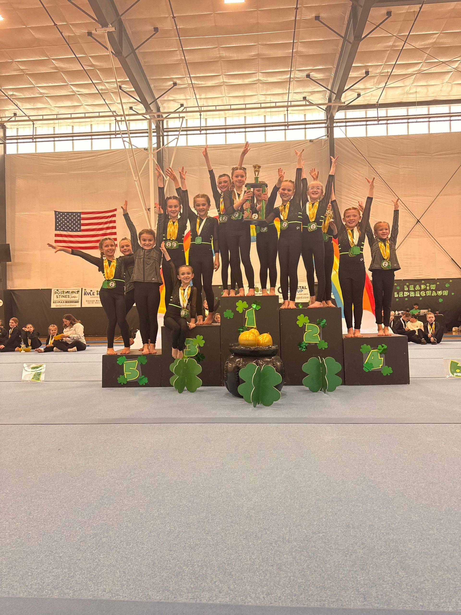 Group of athletes on a podium indoors, raising medals and trophies with an American flag backdrop.