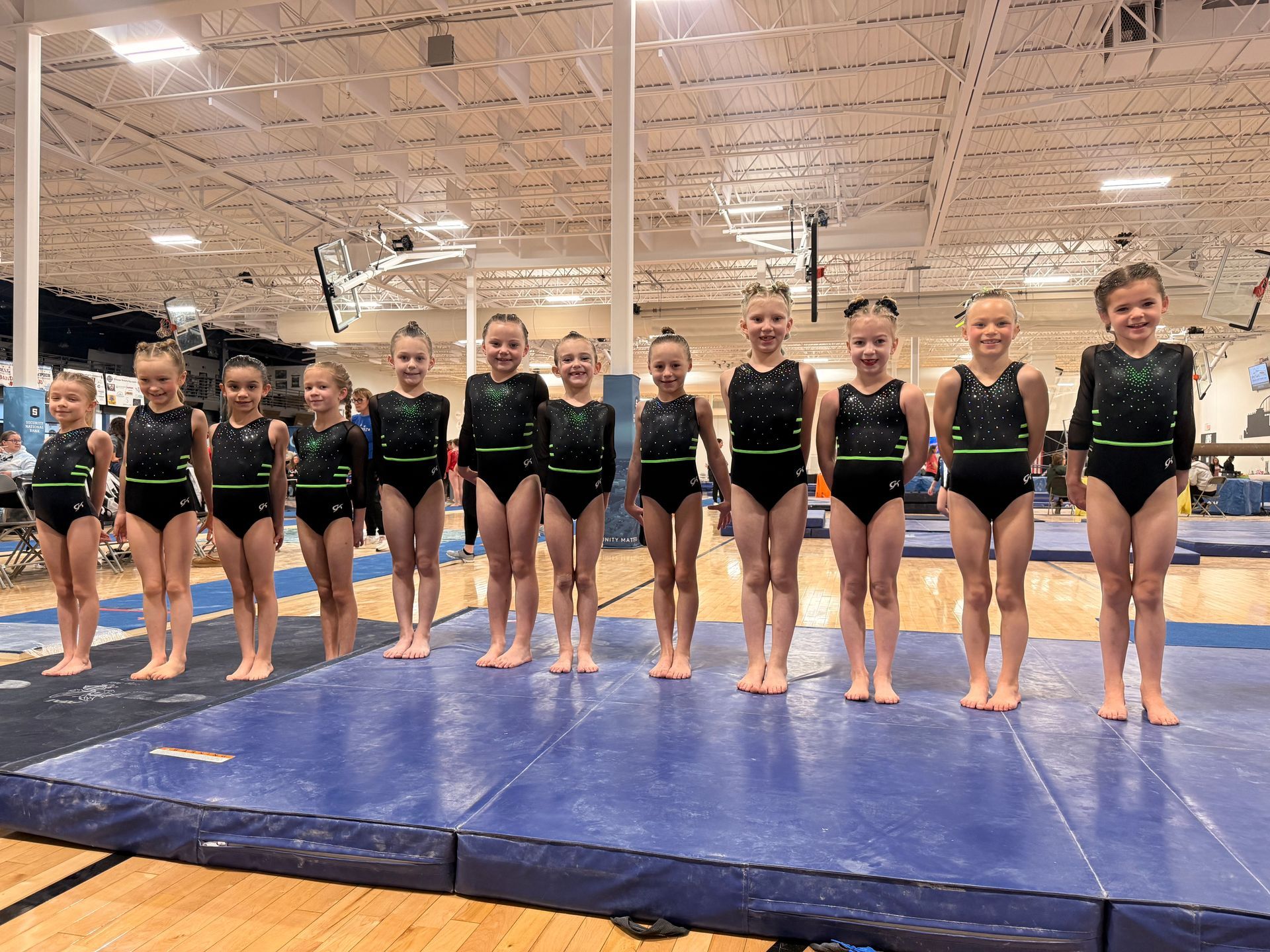 Gymnasts posing in black leotards on blue mats inside a large indoor gym