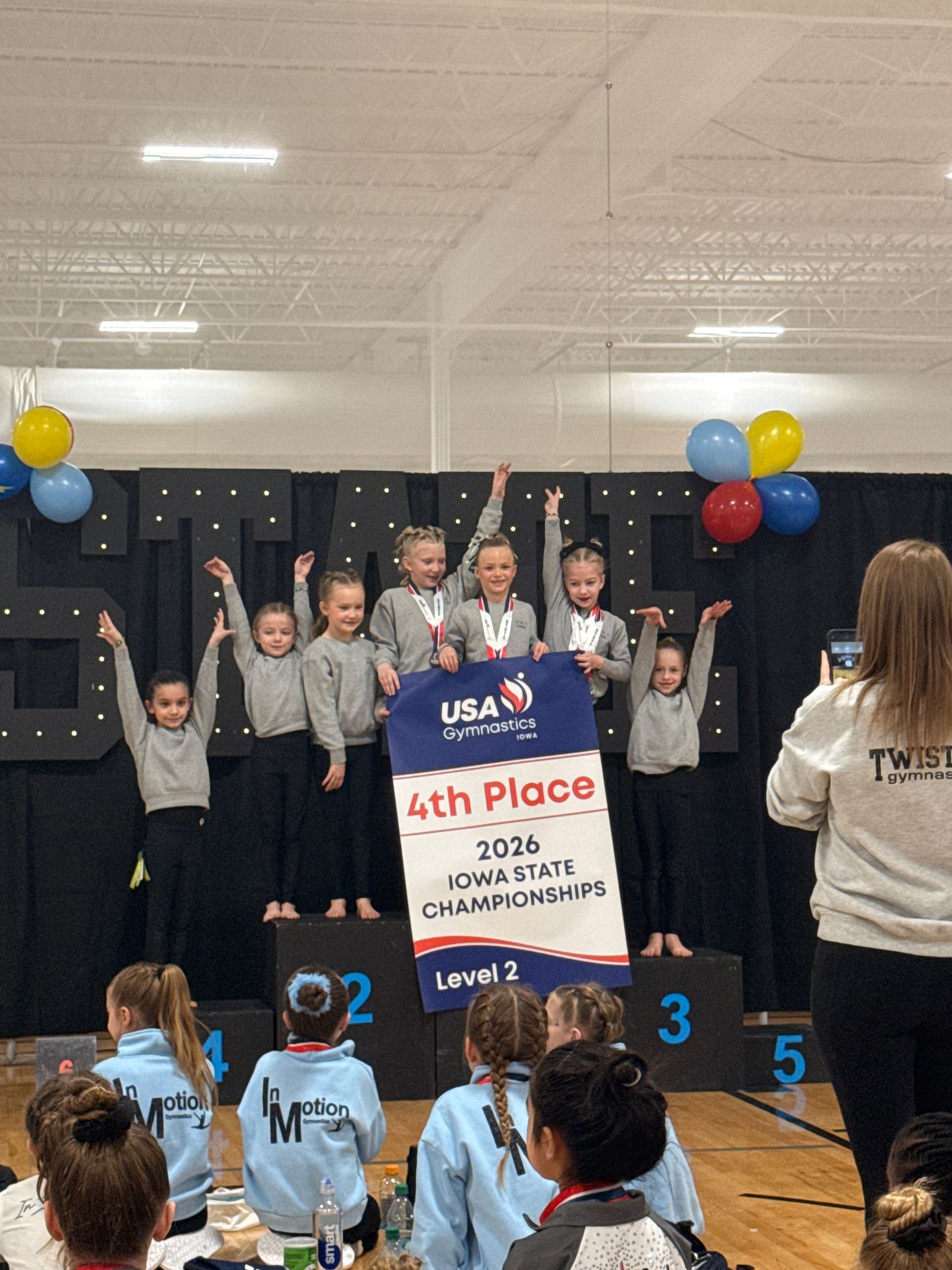 School award ceremony with children on stage holding a 4th place sign and balloons in the background