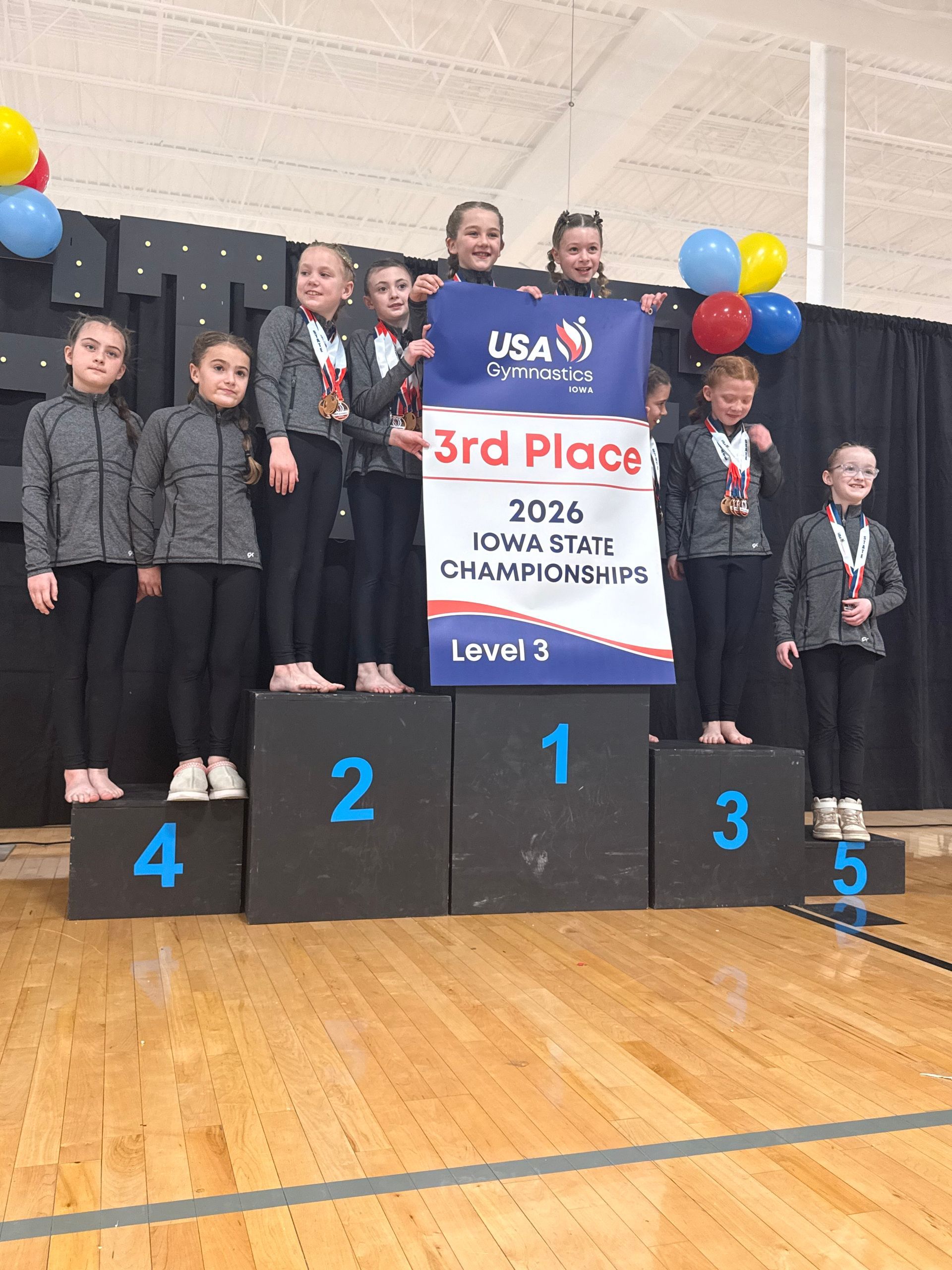 Youth cheer team on podium with 3rd place banner, balloons, and medals at awards ceremony