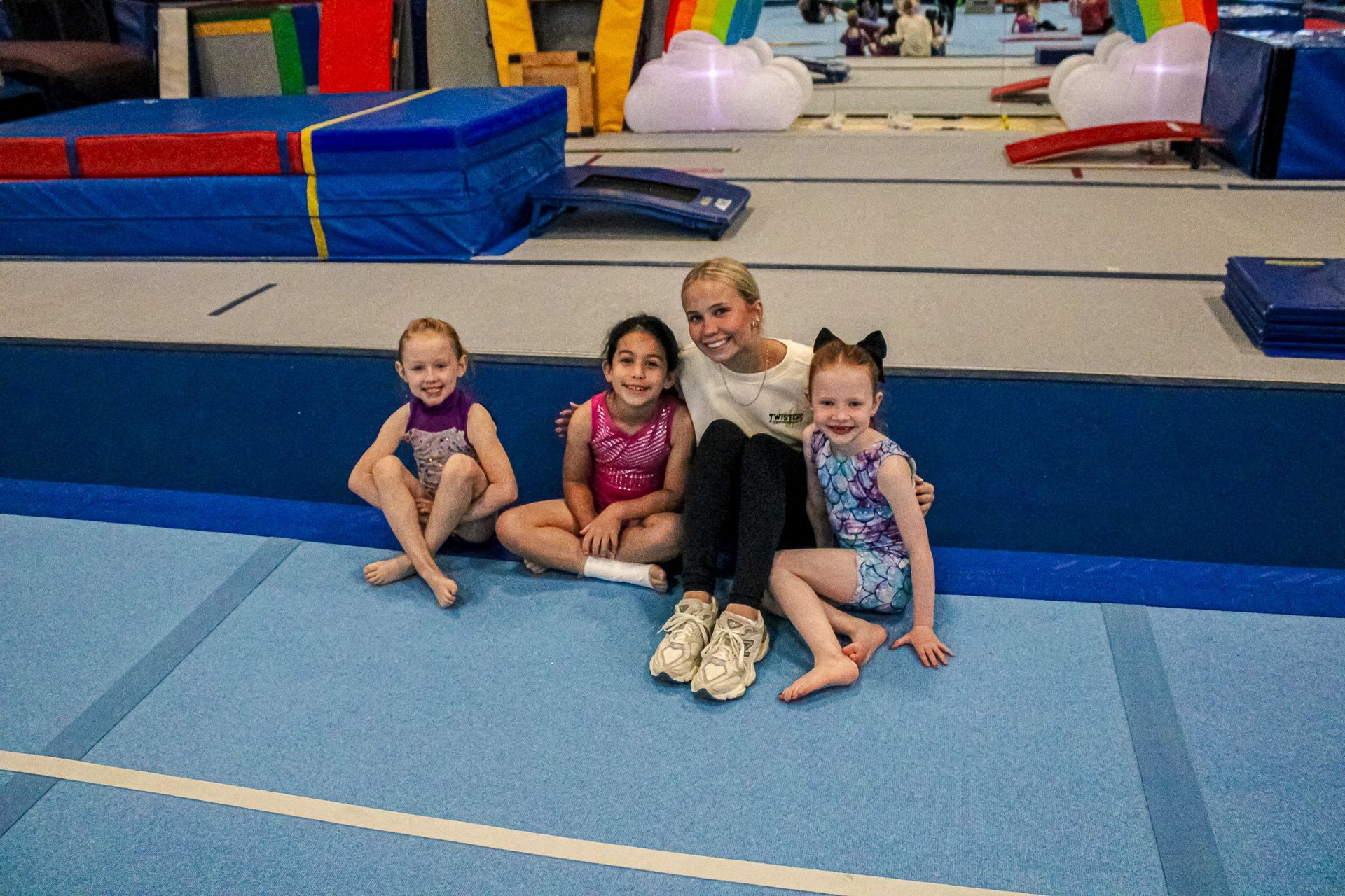Four children sitting on blue gymnastics mats in a gym, with foam equipment in the background.
