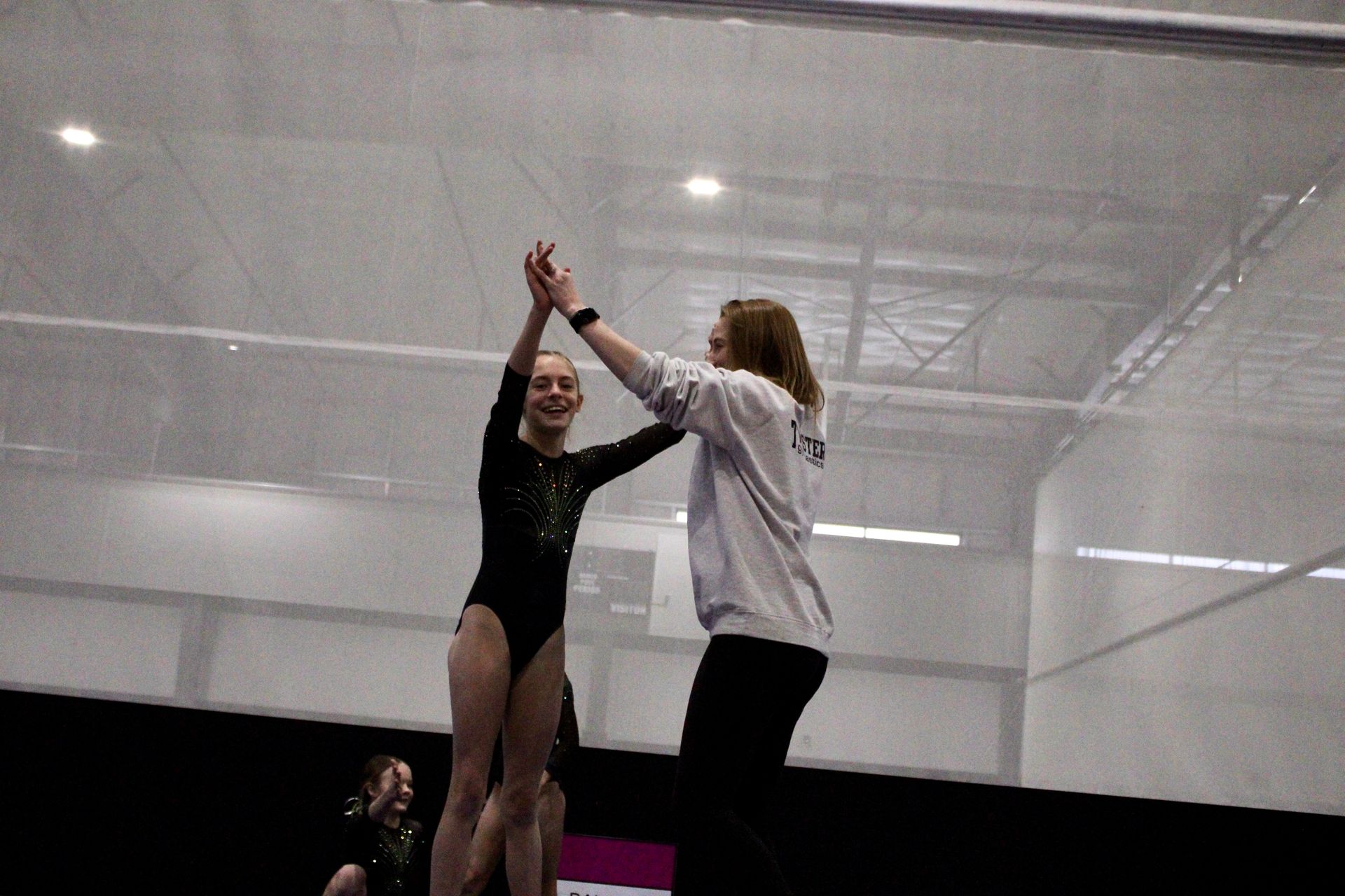 Two performers practicing a lift on a stage under white netting, one in black leotard and one in a gray hoodie
