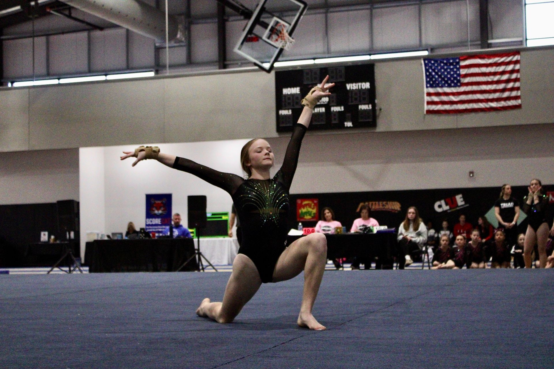 Gymnast kneeling on a blue mat with arms extended, performing under gym lights with spectators and U.S. flag in background