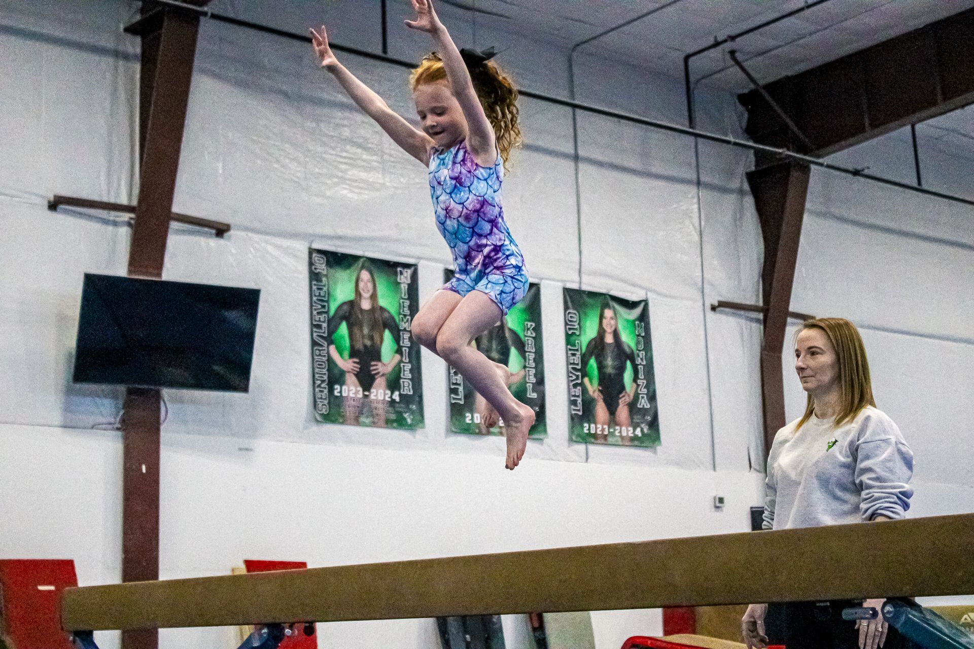 Gymnast on a balance beam in a gym, with a coach watching nearby.
