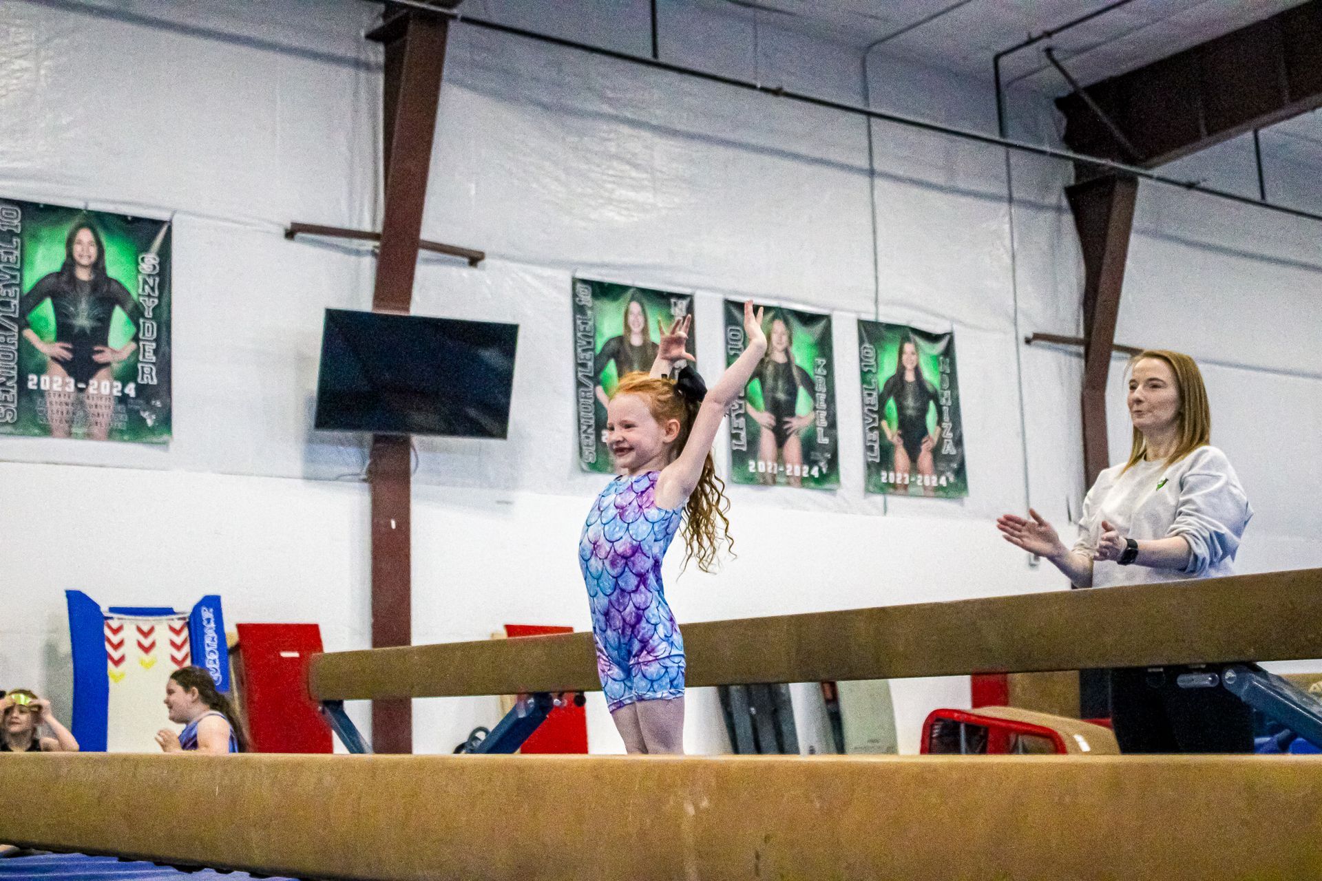 Child in a colorful leotard raises her arms in a gym, with a coach and banners in the background.