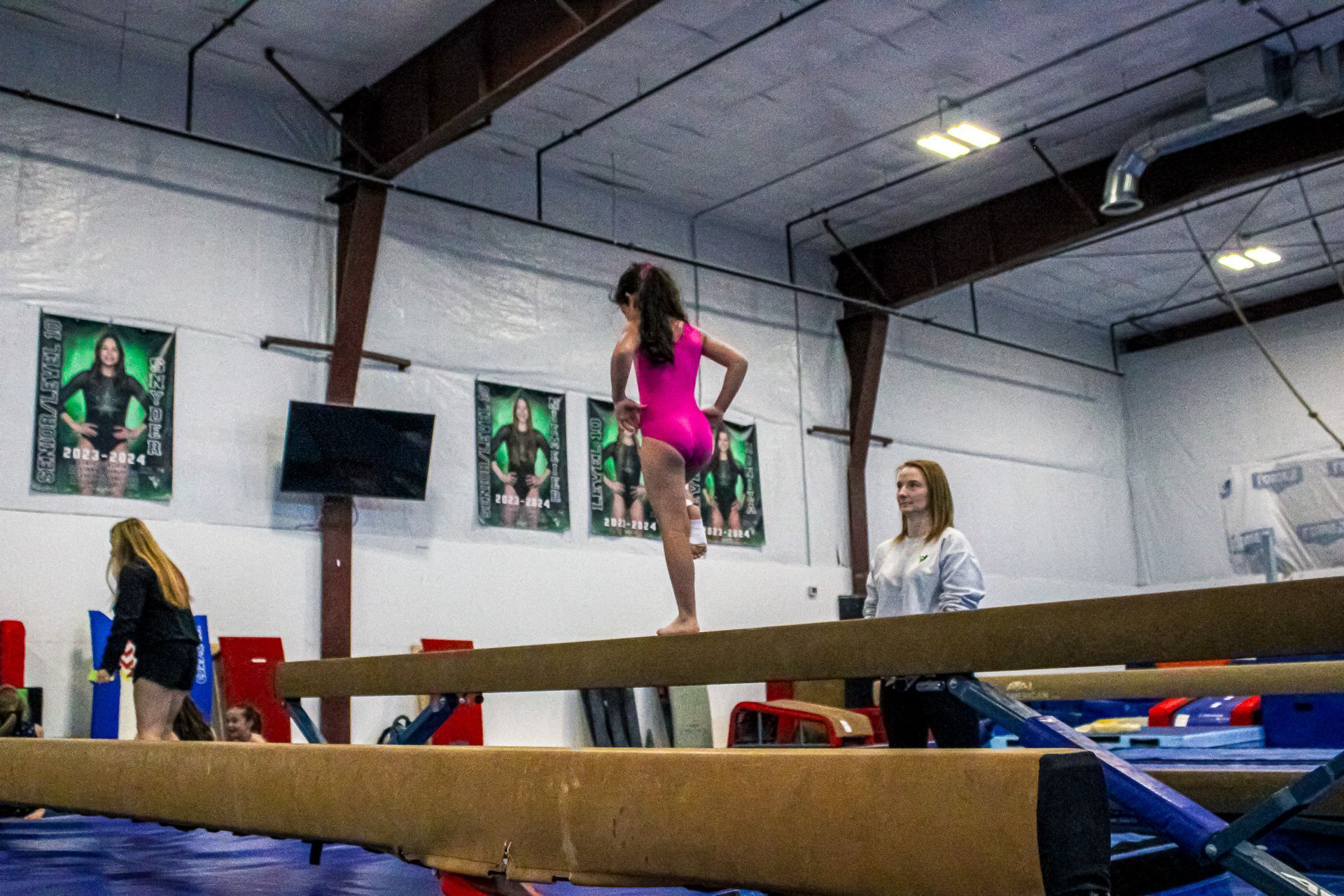 Gymnast balancing on a beam in an indoor gym, with coaches nearby.