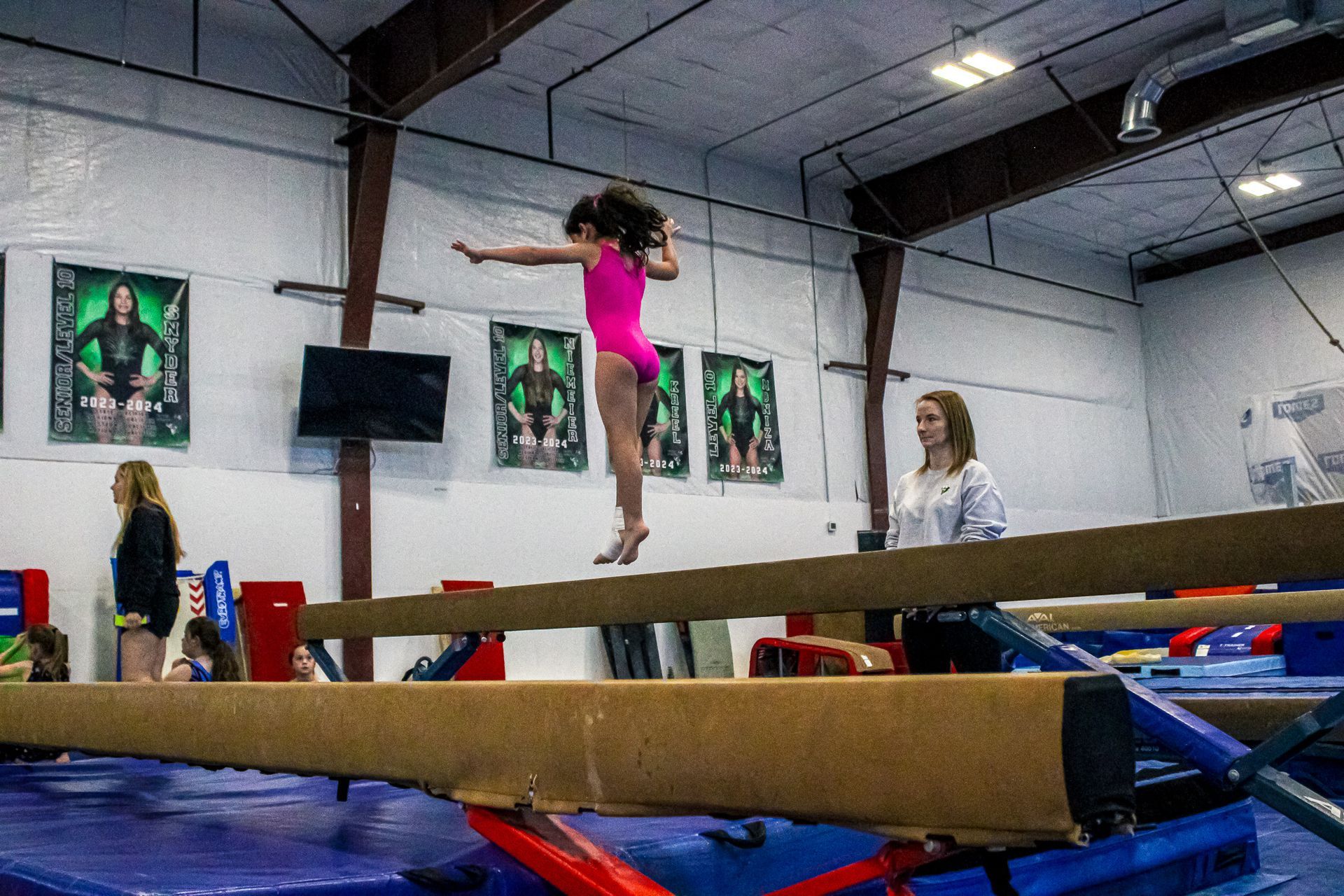 Gymnast balancing on a beam in a training gym with coaches and mats nearby