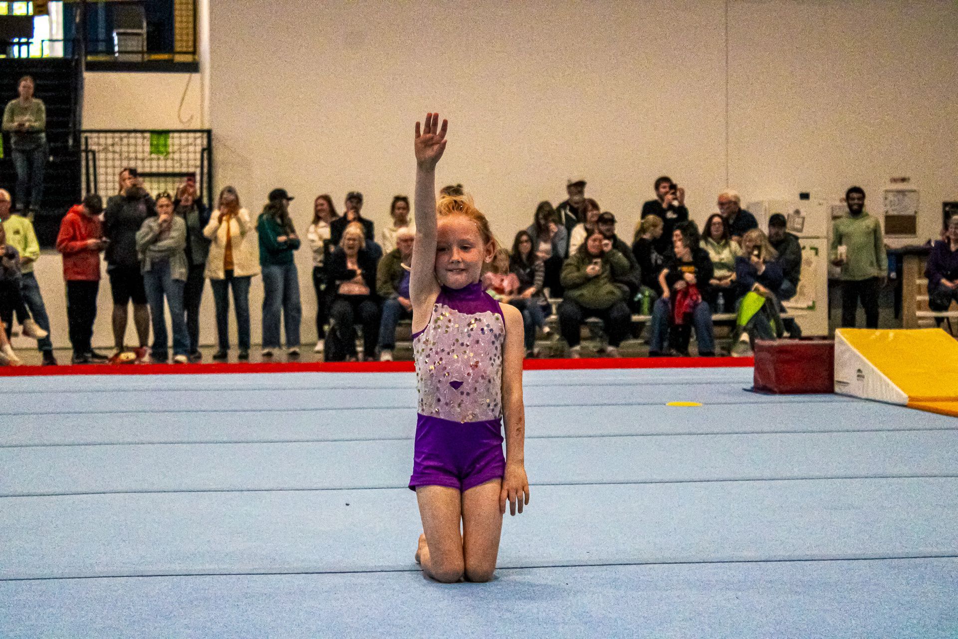 Gymnast kneeling on a blue mat, one arm raised, with spectators watching in the background.