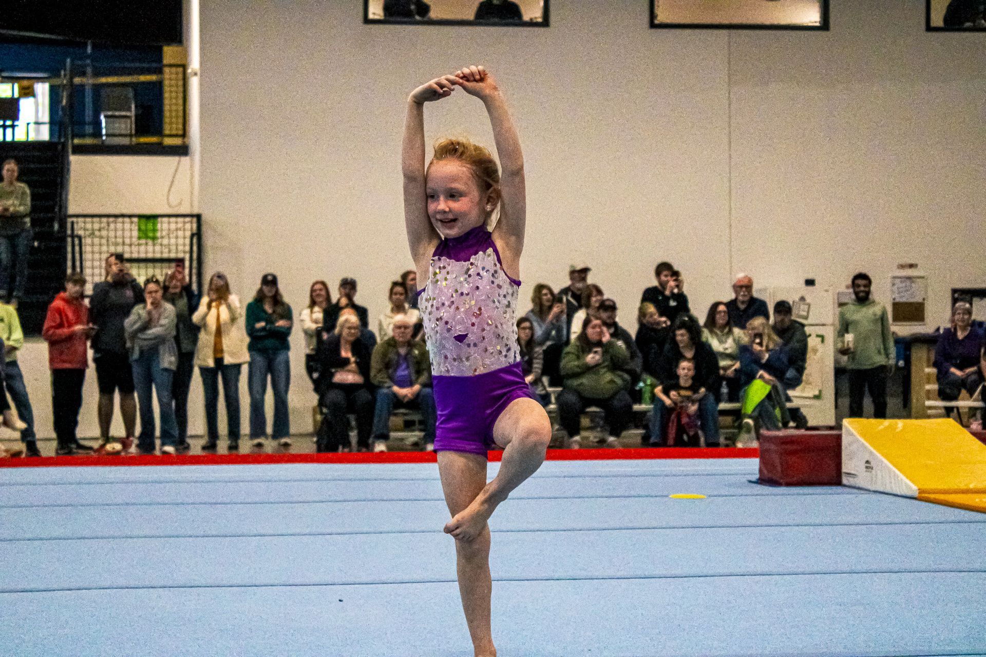 Young gymnast in purple leotard balancing in a dance pose on a blue floor mat, spectators behind her