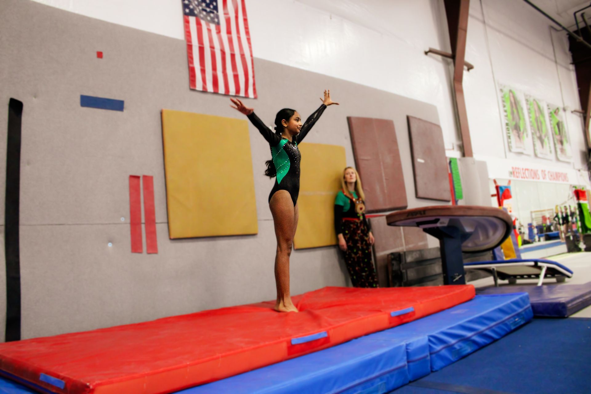 Gymnast balancing on a beam in a gym, arms raised, with mats and an American flag in the background.