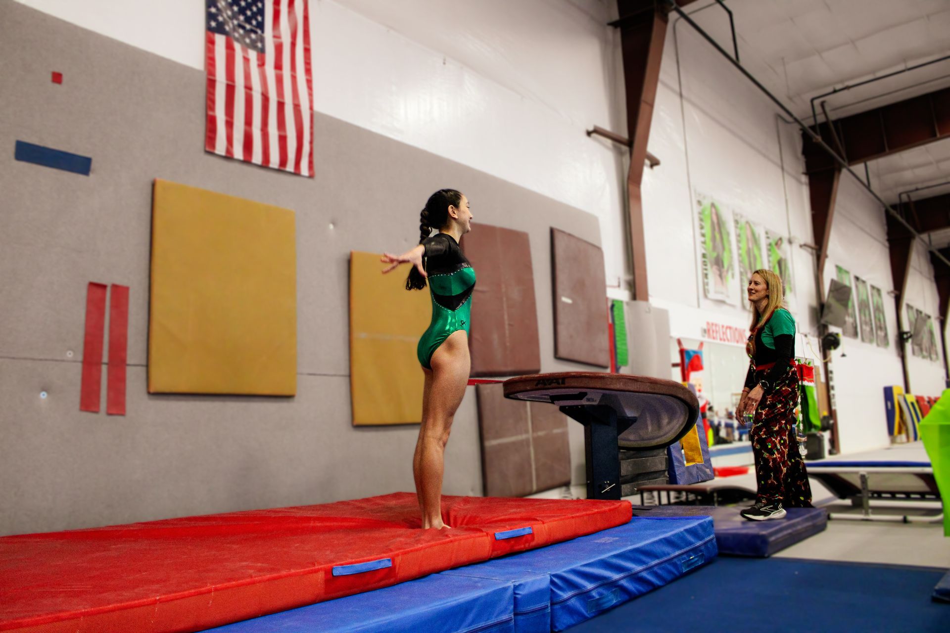 Two gymnasts in leotards stand on mats in a gymnastics gym with an American flag on the wall