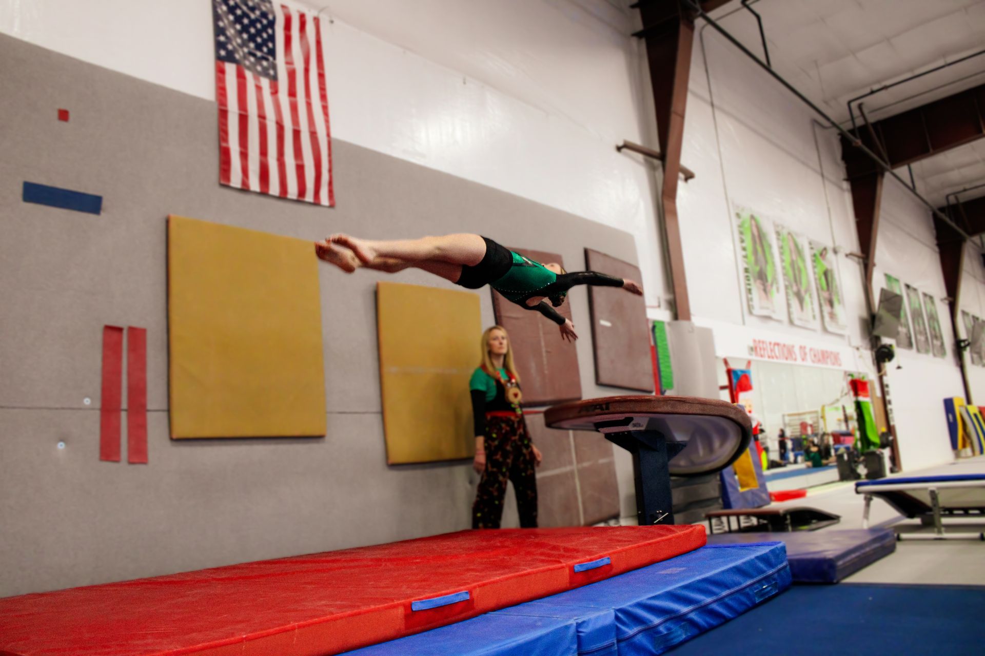 Gymnast balancing horizontally on bars in an indoor gym with red and blue mats and an American flag.