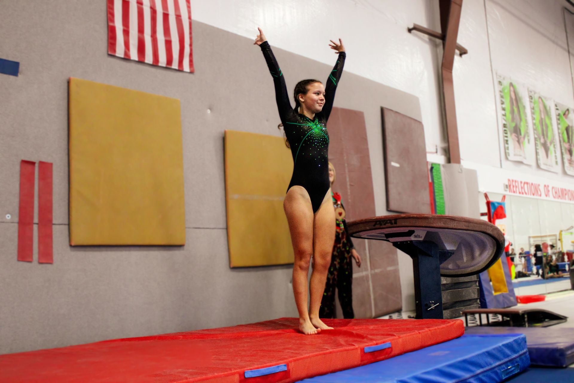 Gymnast in black leotard standing on a red mat with arms raised in a gym.