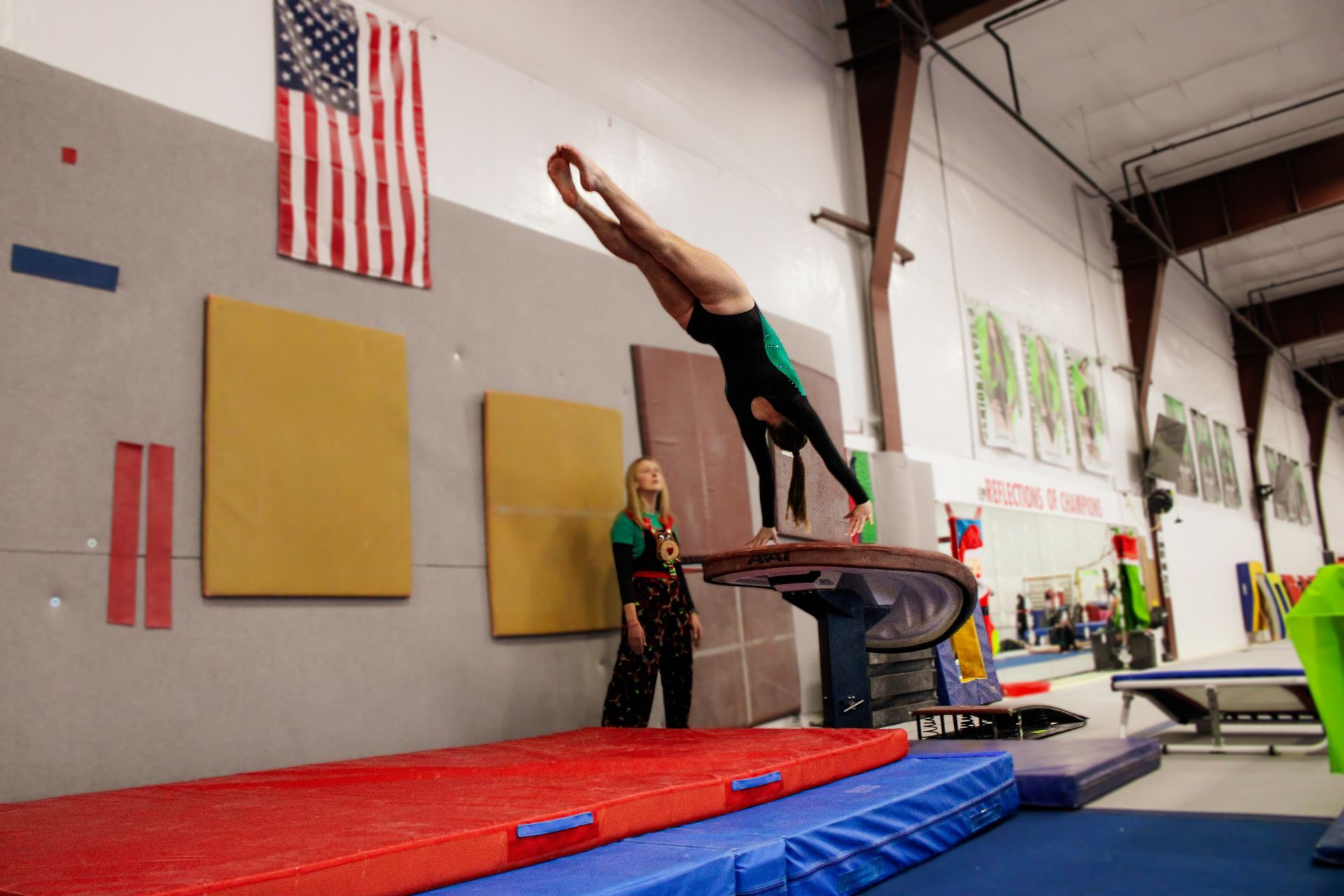 Gymnast vaulting over a padded table in an indoor gym, with a coach nearby and an American flag on the wall
